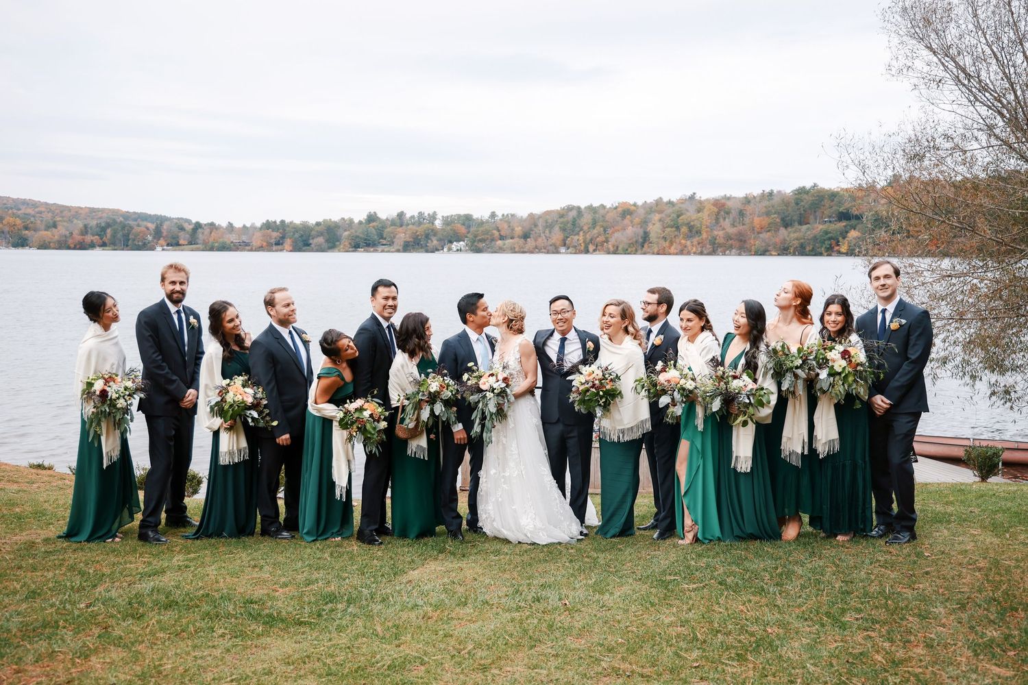 Wedding party in emerald green dresses and dark suits pose together by a scenic autumn lake during golden hour.