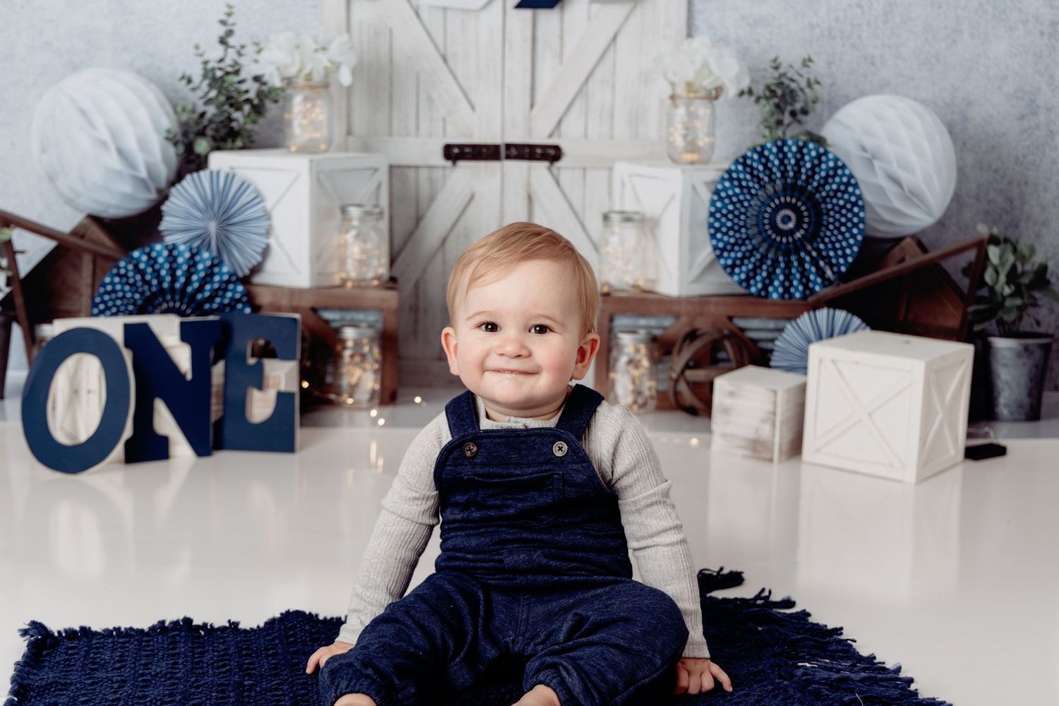 A baby wearing a navy blue winter outfit sits on a white blanket with holiday decorations in the background.