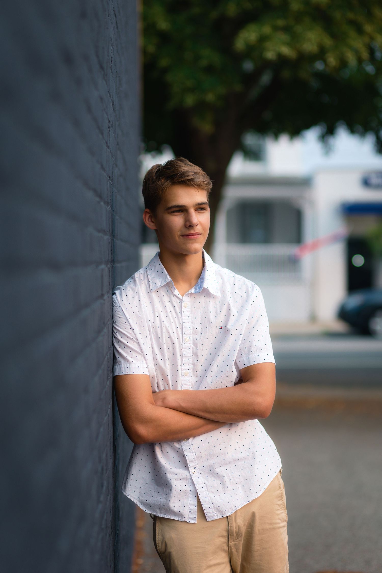 A person in a white patterned shirt and khaki pants leans against a gray wall in an outdoor setting.