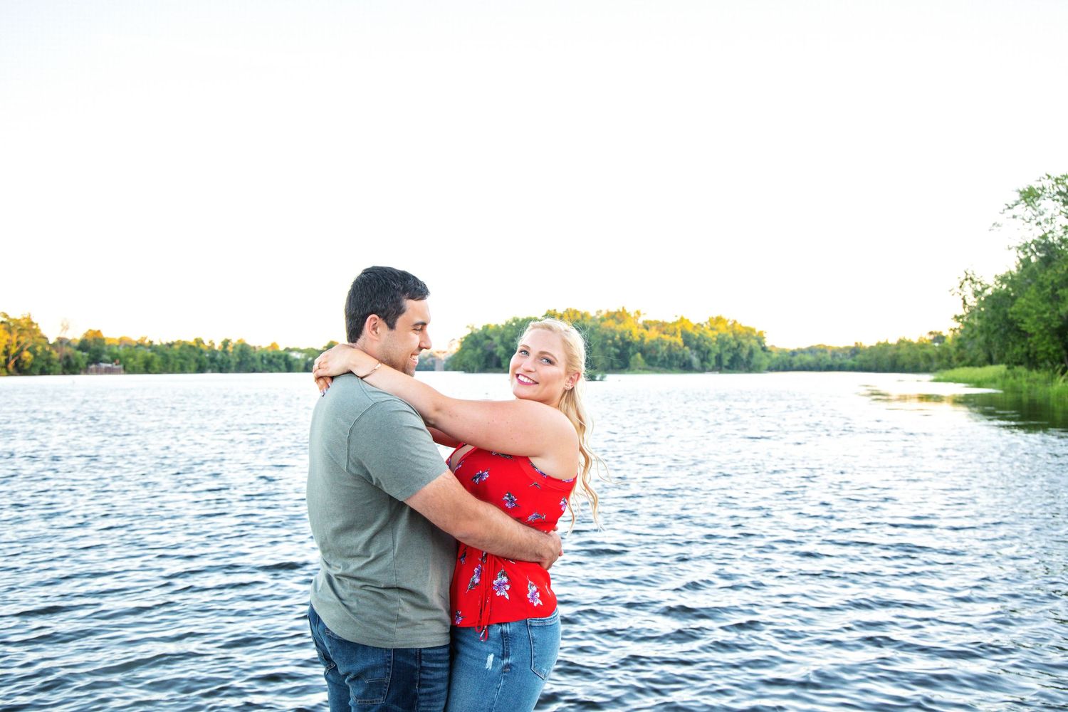 A couple embraces near a serene lake with sunflowers in the background during a summer engagement photo session.