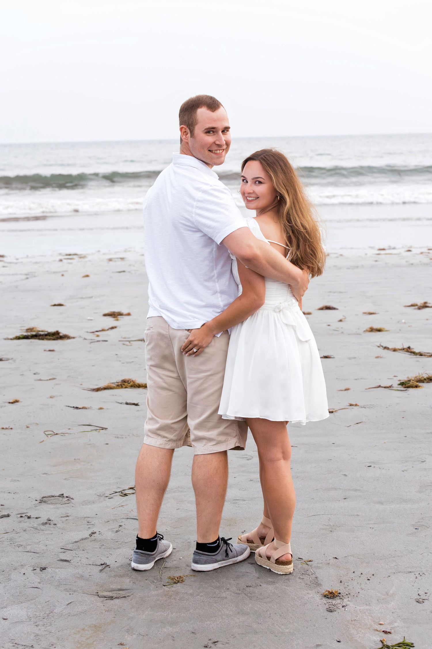 A couple in white and khaki outfits shares playful moments on a beach with seaweed and waves in the background.