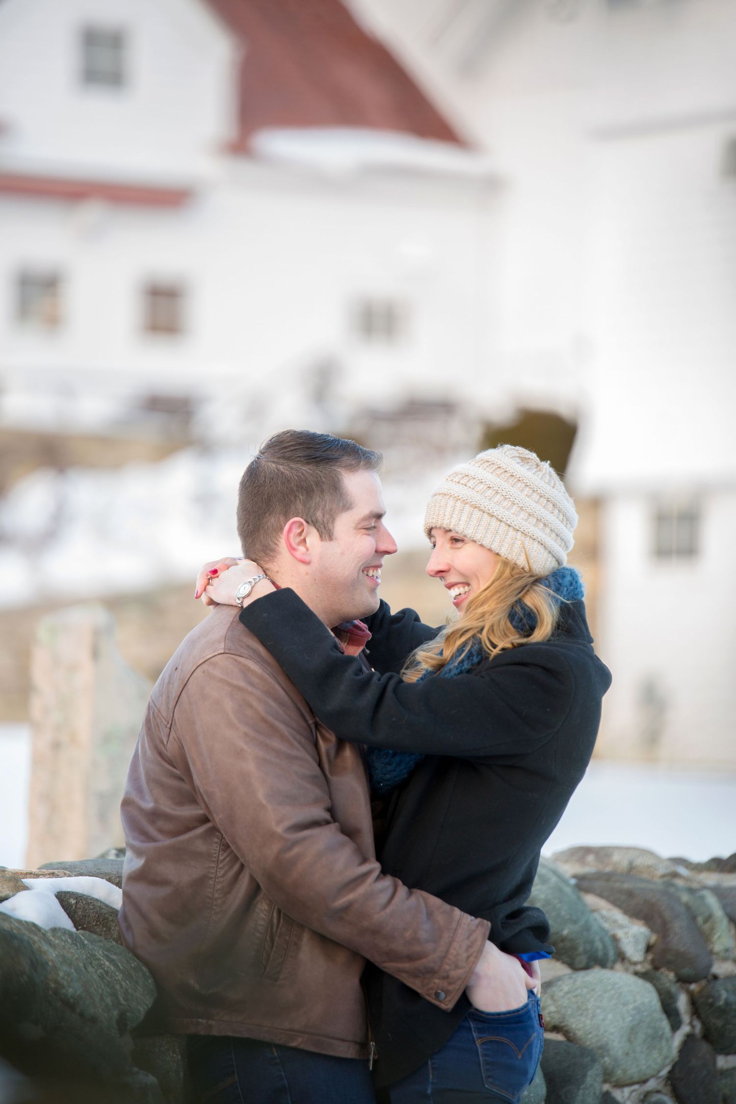 A couple sits together and laughs on a stone wall during a winter engagement photoshoot.
