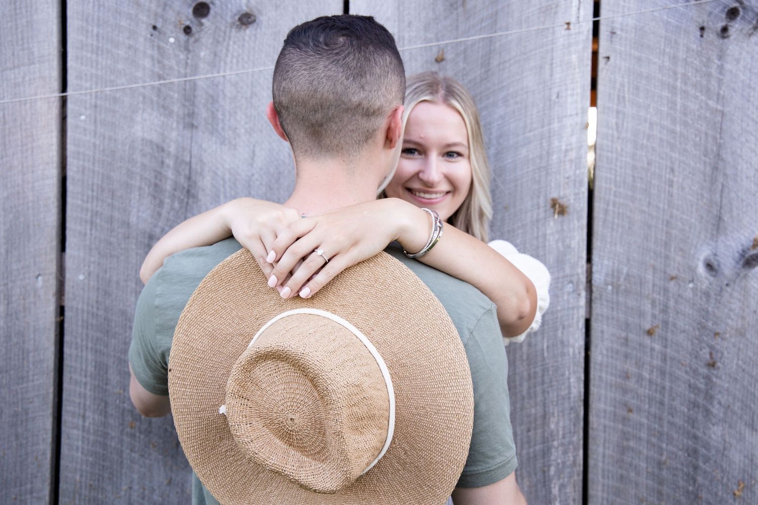 A couple embraces against a rustic wooden wall, with a tan straw hat visible in the foreground.
