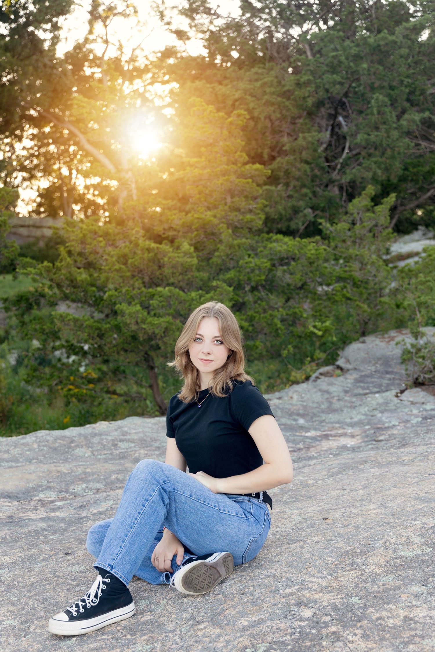 A person in a black shirt and jeans reclines on a rocky outcrop during golden hour sunset in a wooded area.