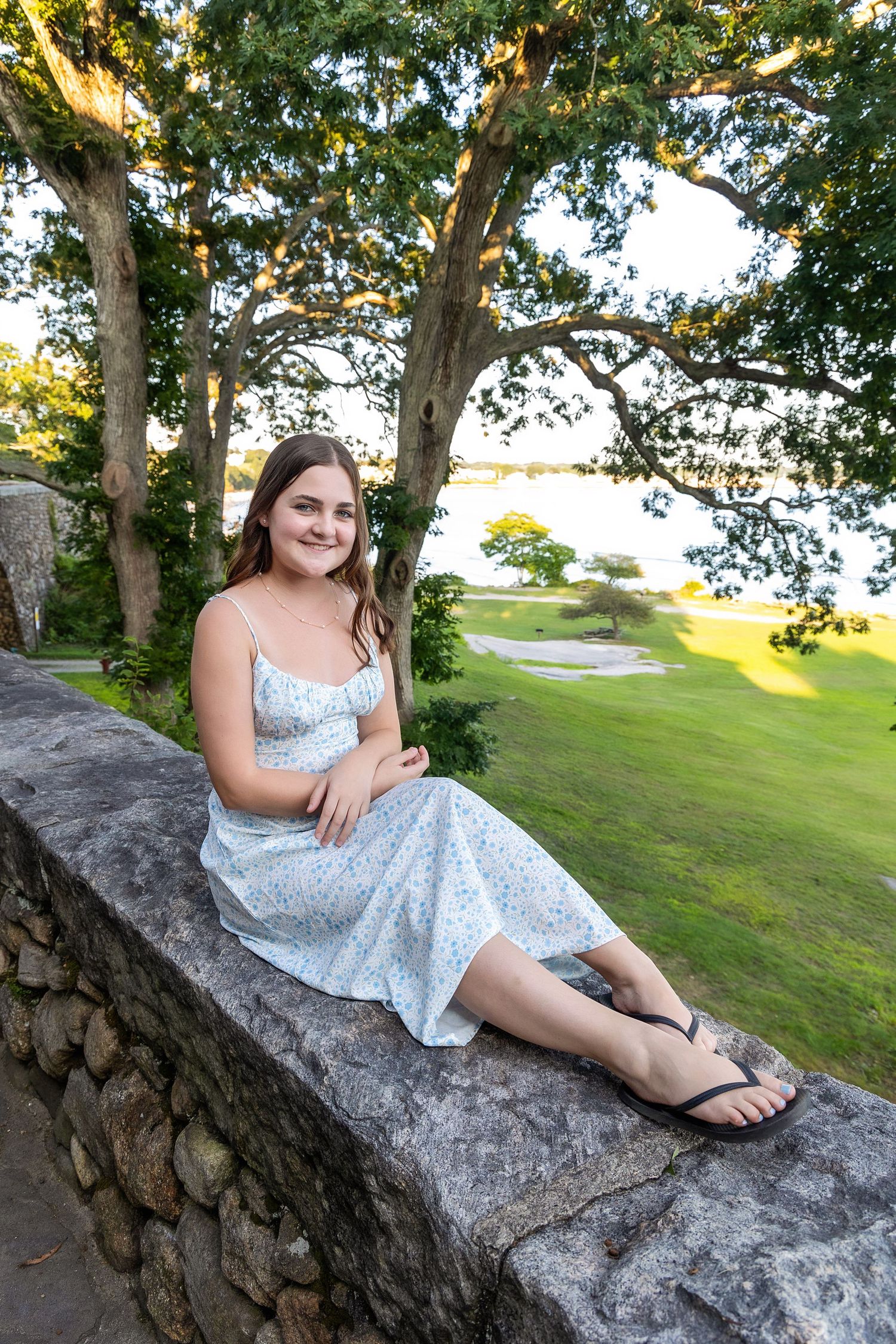 A person in a light blue floral dress relaxes on a stone wall surrounded by green trees and grass.