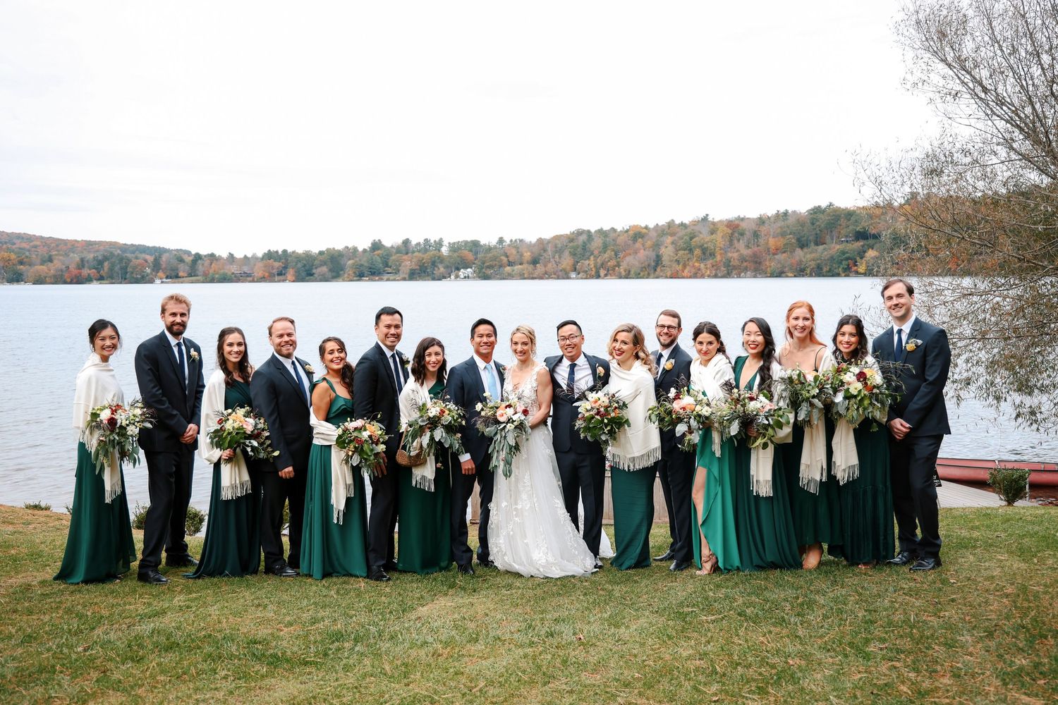 Wedding party in green dresses and dark suits poses outdoors by a scenic lake on an autumn day.