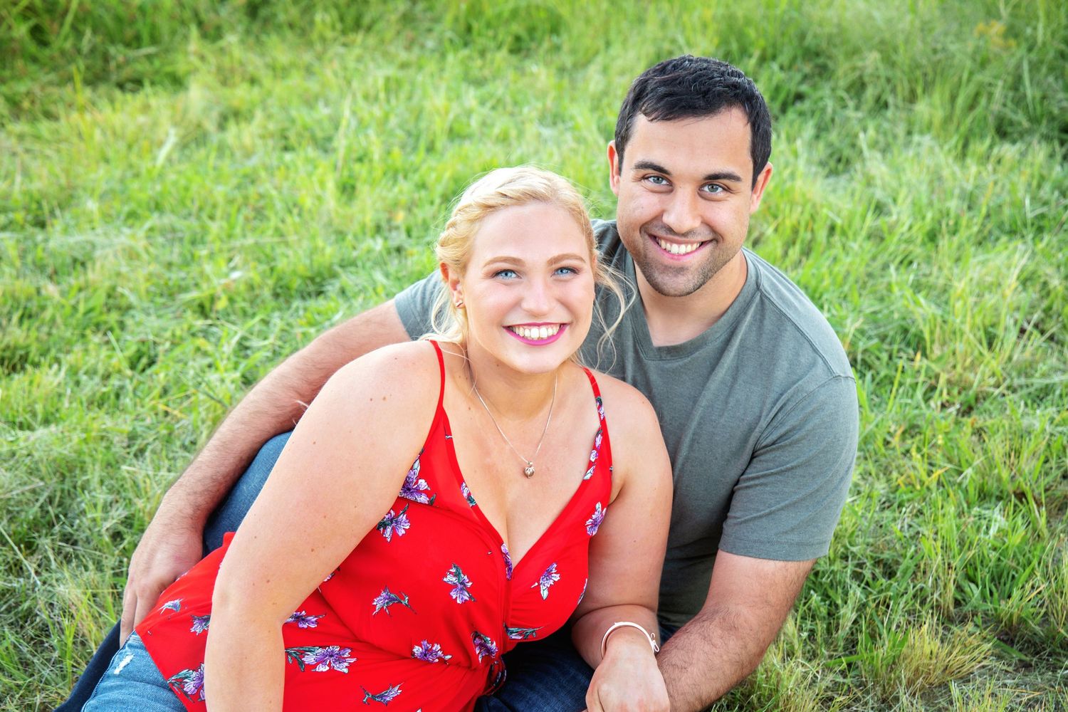 A couple sits together outdoors on green grass wearing red and grey casual clothing on a sunny day.