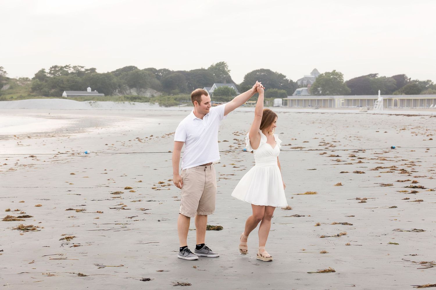 A couple in white outfits walking hand in hand along a scenic beach at sunset.