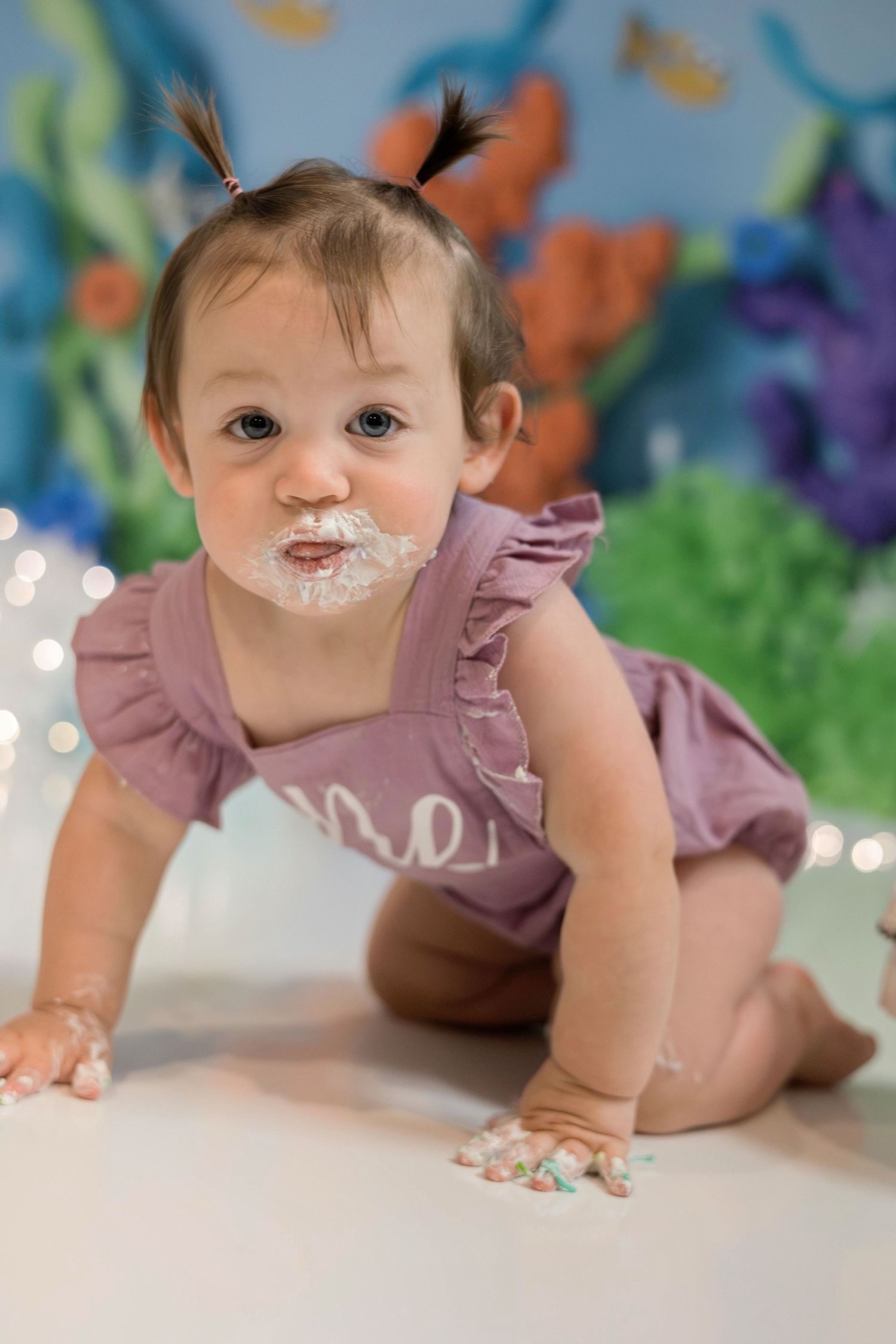 Baby in dusty pink ruffled outfit with cake frosting on face crawls on floor during colorful birthday photo session.