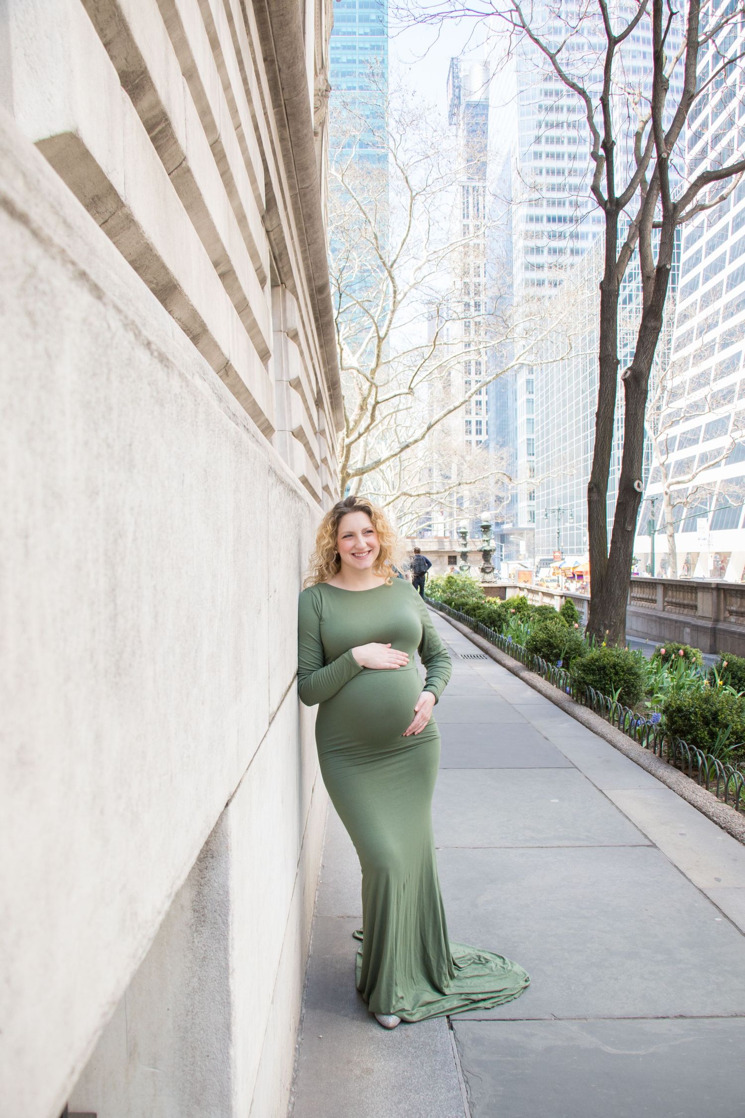 A pregnant woman in a flowing olive green dress poses against a modern urban backdrop with concrete columns and trees.