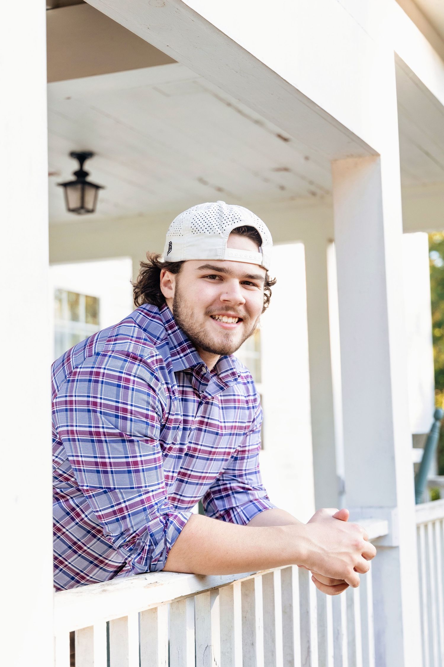 Person in plaid shirt and white cap smiles while leaning on white porch columns on a sunny day.