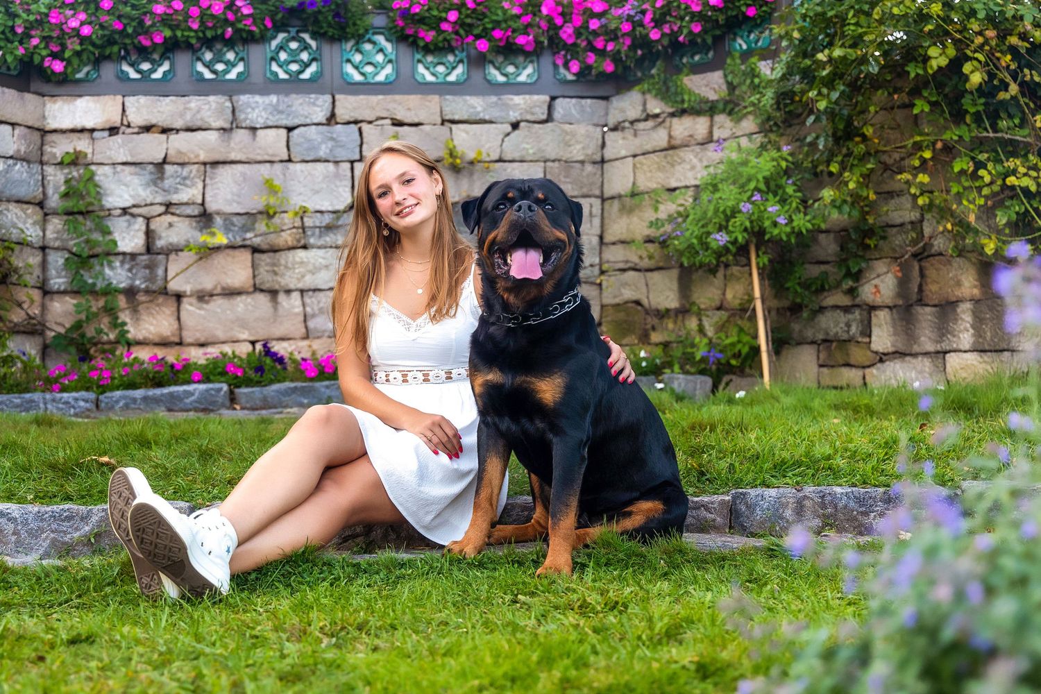 A person and black dog relax together on green grass near a stone wall with colorful flowers blooming nearby.