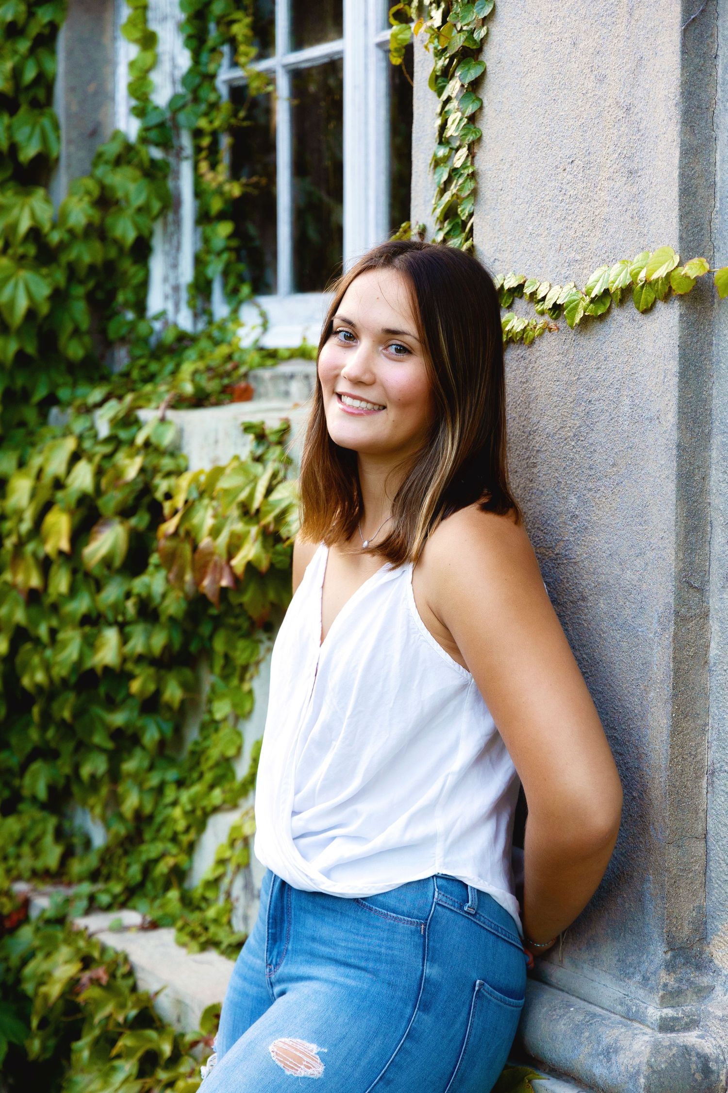 A person in a white sleeveless top and blue jeans poses against a stone wall with ivy growing in the background.