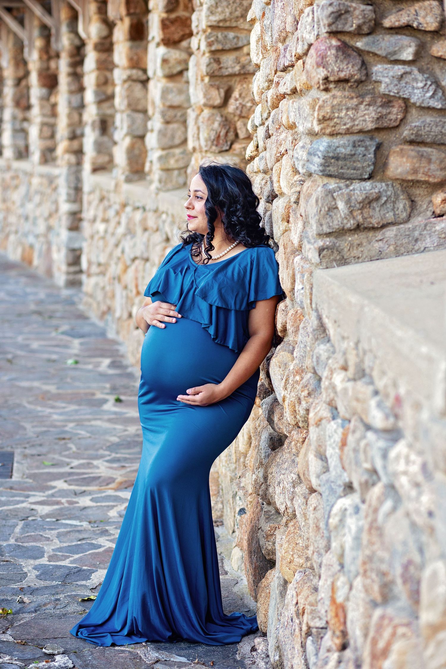 A woman in a royal blue maternity dress poses against a rustic stone wall for pregnancy photos.