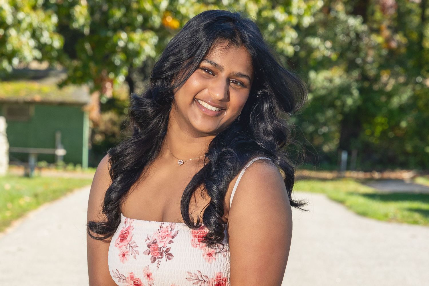 A person in a floral dress poses outdoors on a sunny day with trees and greenery in the background.