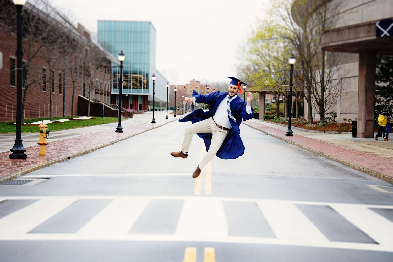 A skateboarder performs a trick on a long concrete walkway surrounded by trees and buildings on a college campus.