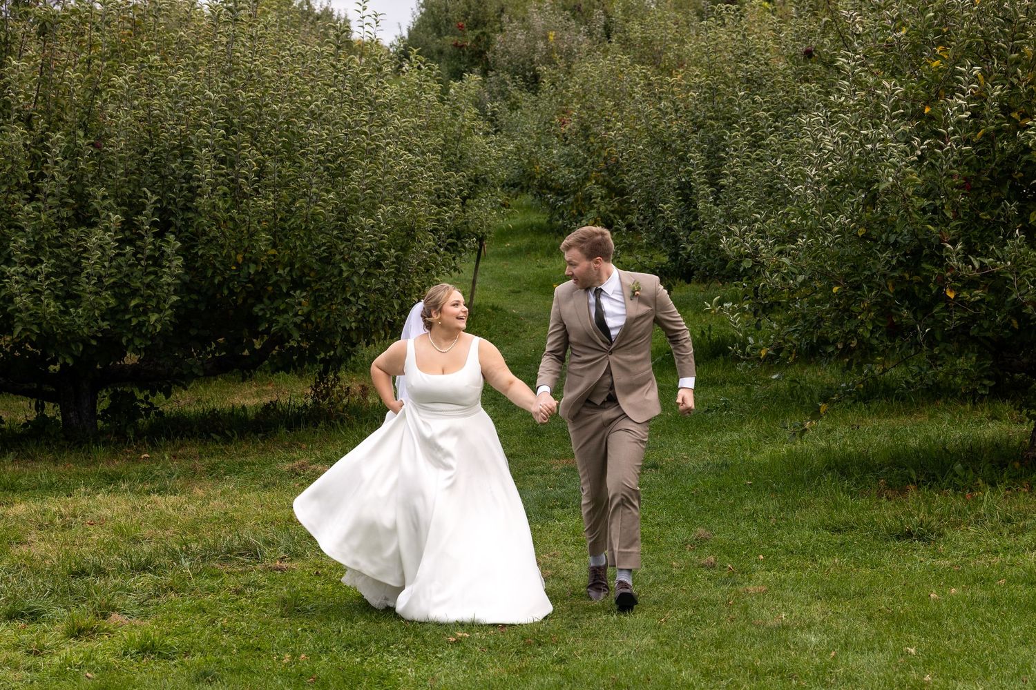 Newlyweds run playfully through an apple orchard holding hands.