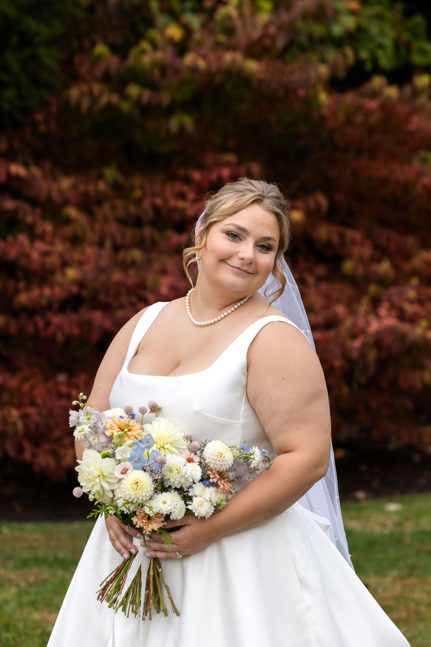 Bride in elegant white dress holds pastel bouquet against fall foliage background.