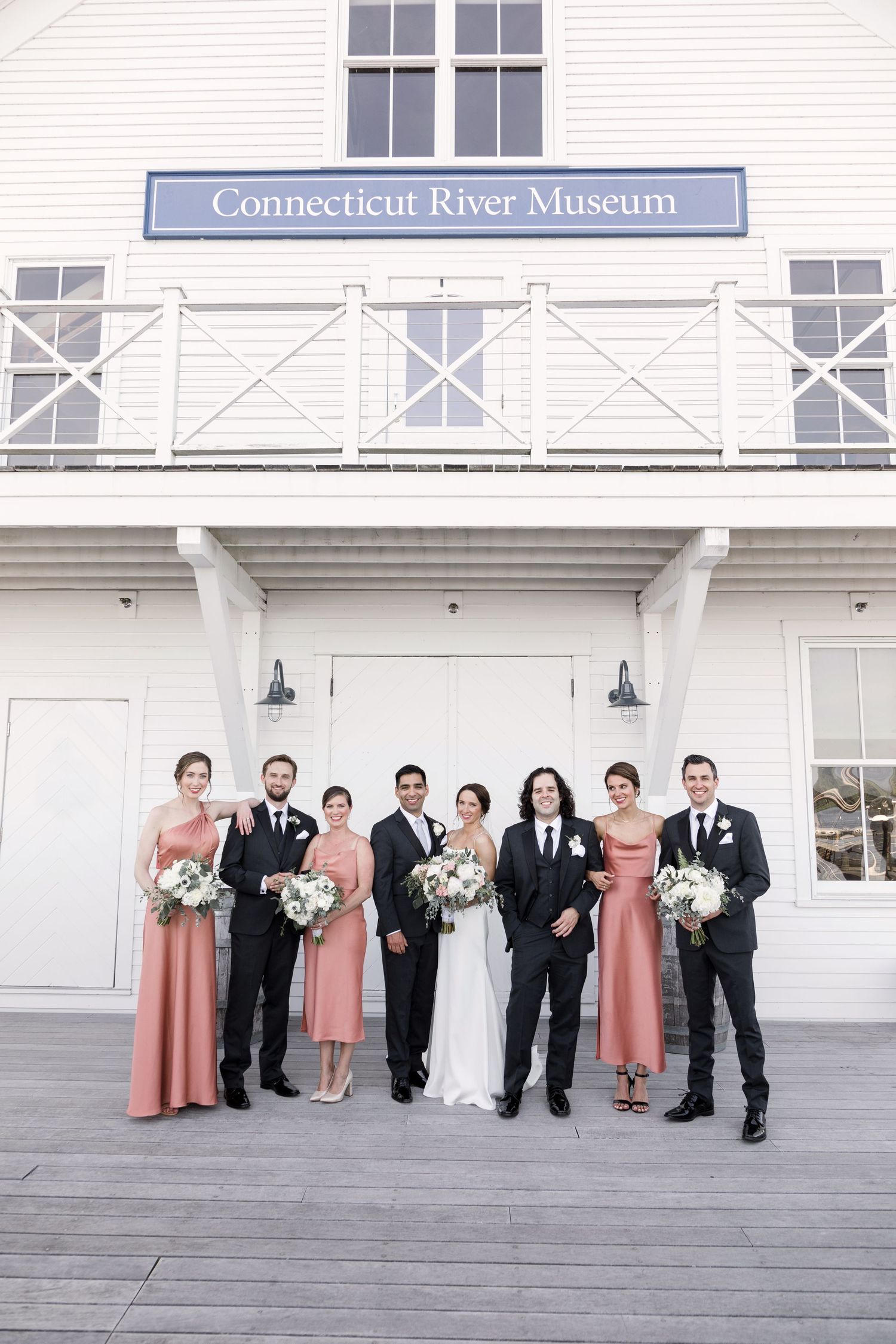 Wedding party poses on wooden deck outside Connecticut River Museum in formal attire with pink bridesmaid dresses.