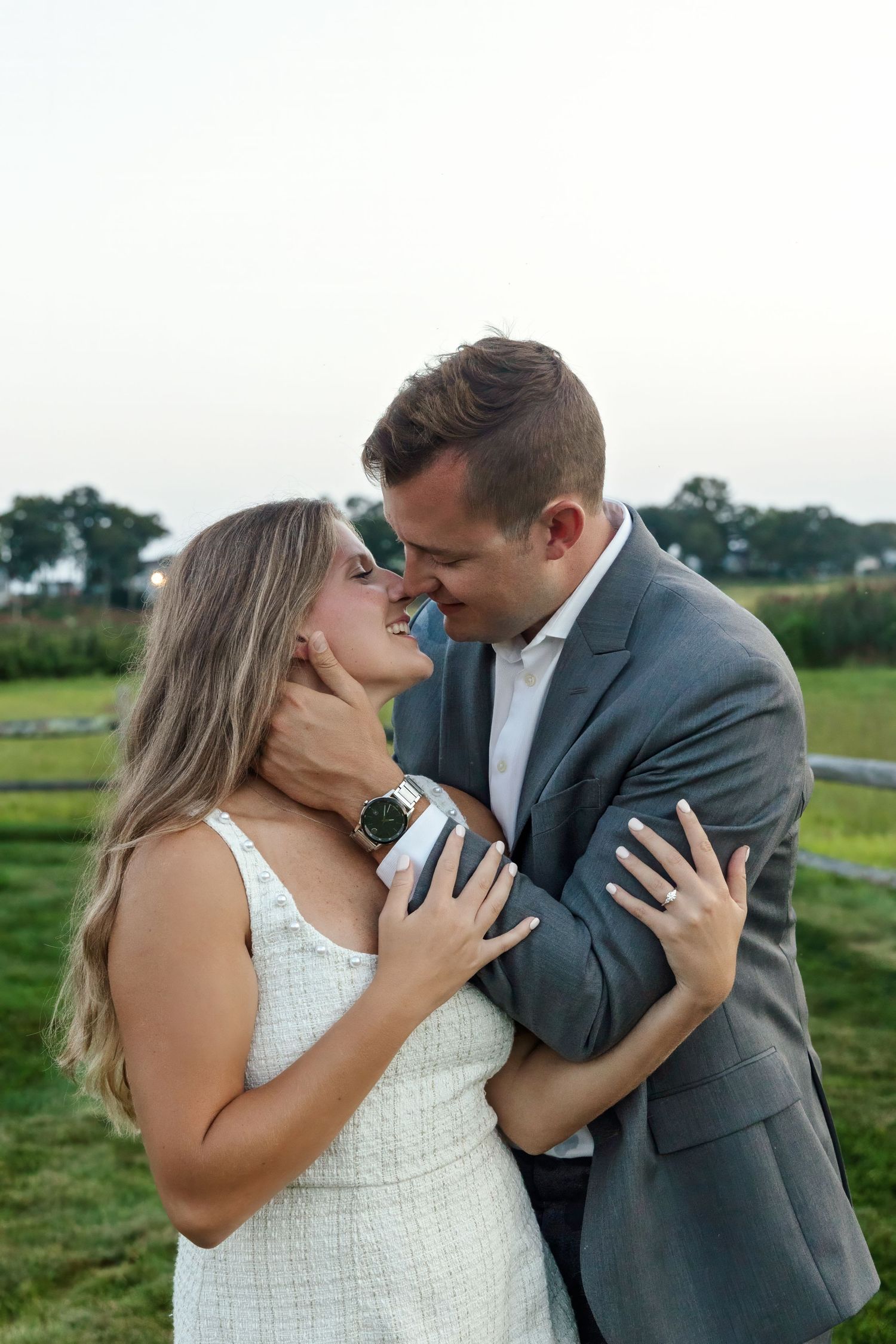 A romantic couple in formal attire share tender kisses in a grassy outdoor setting at sunset.