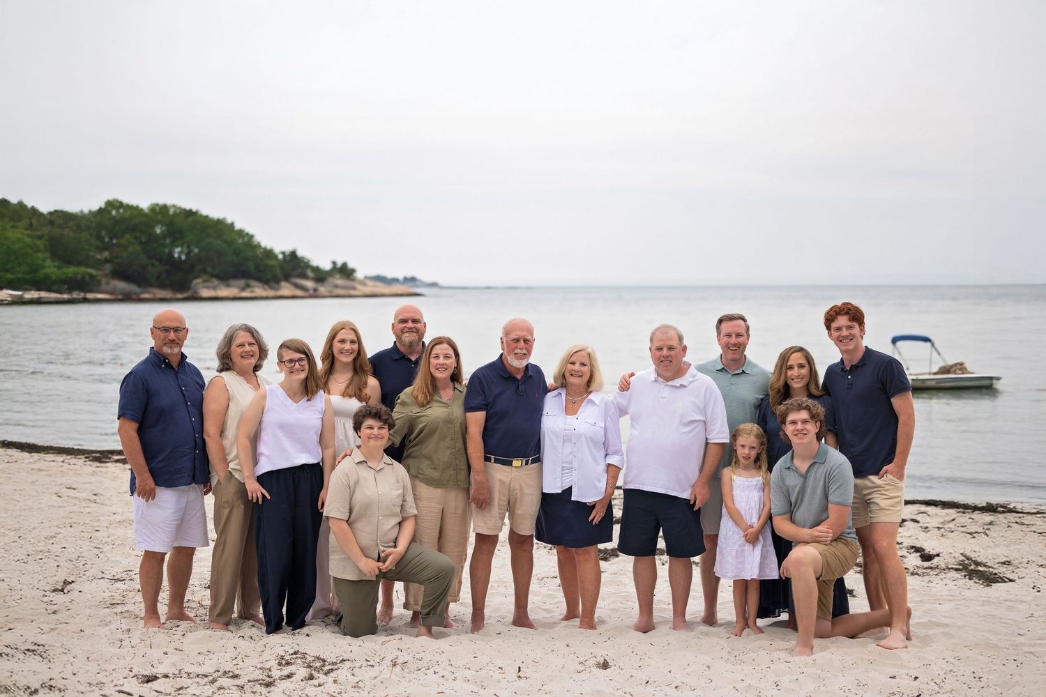 A group of people stand together on a beach with boats and water visible in the background on a cloudy day.