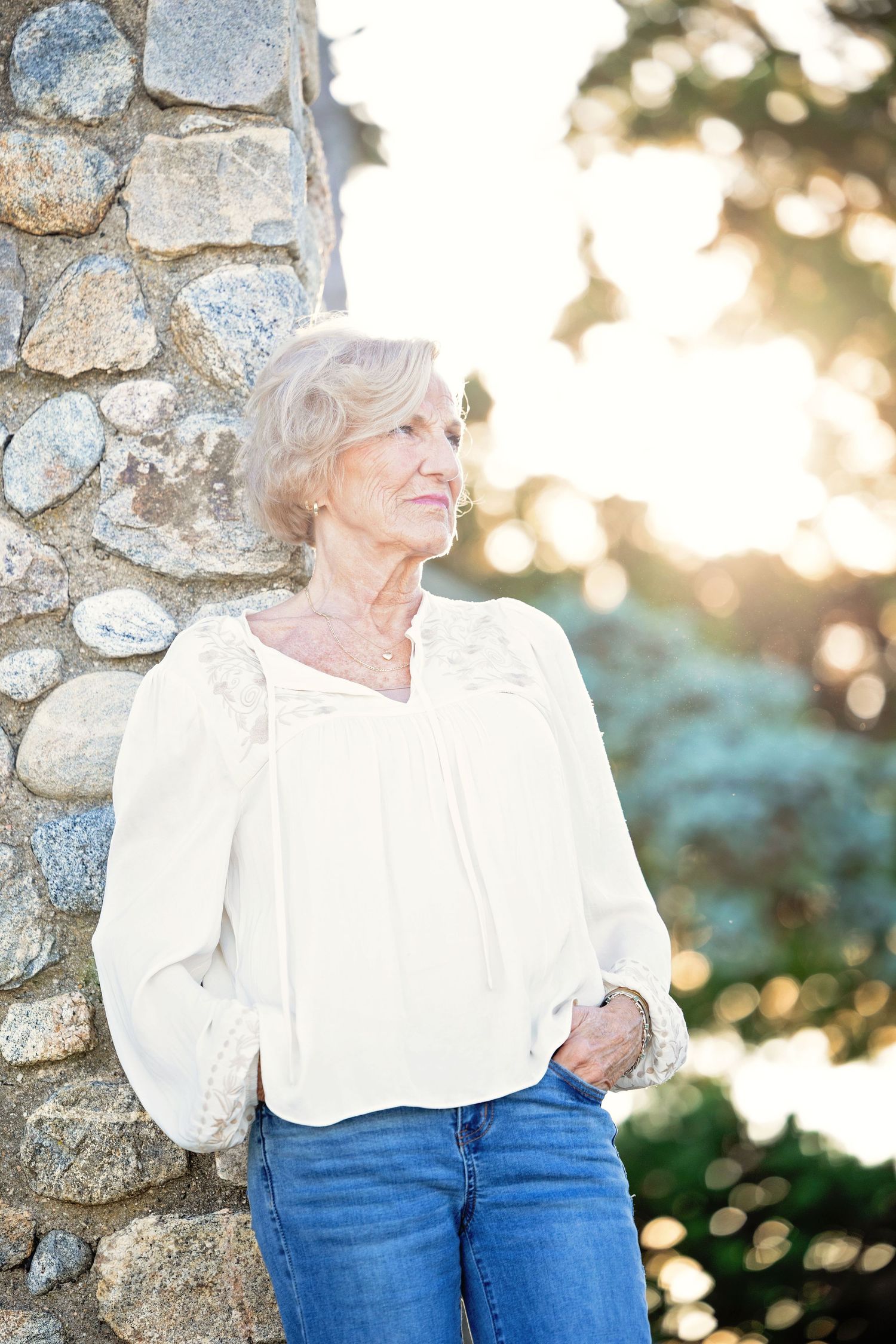 A person in a white blouse and blue jeans leans against a stone wall during golden hour lighting.