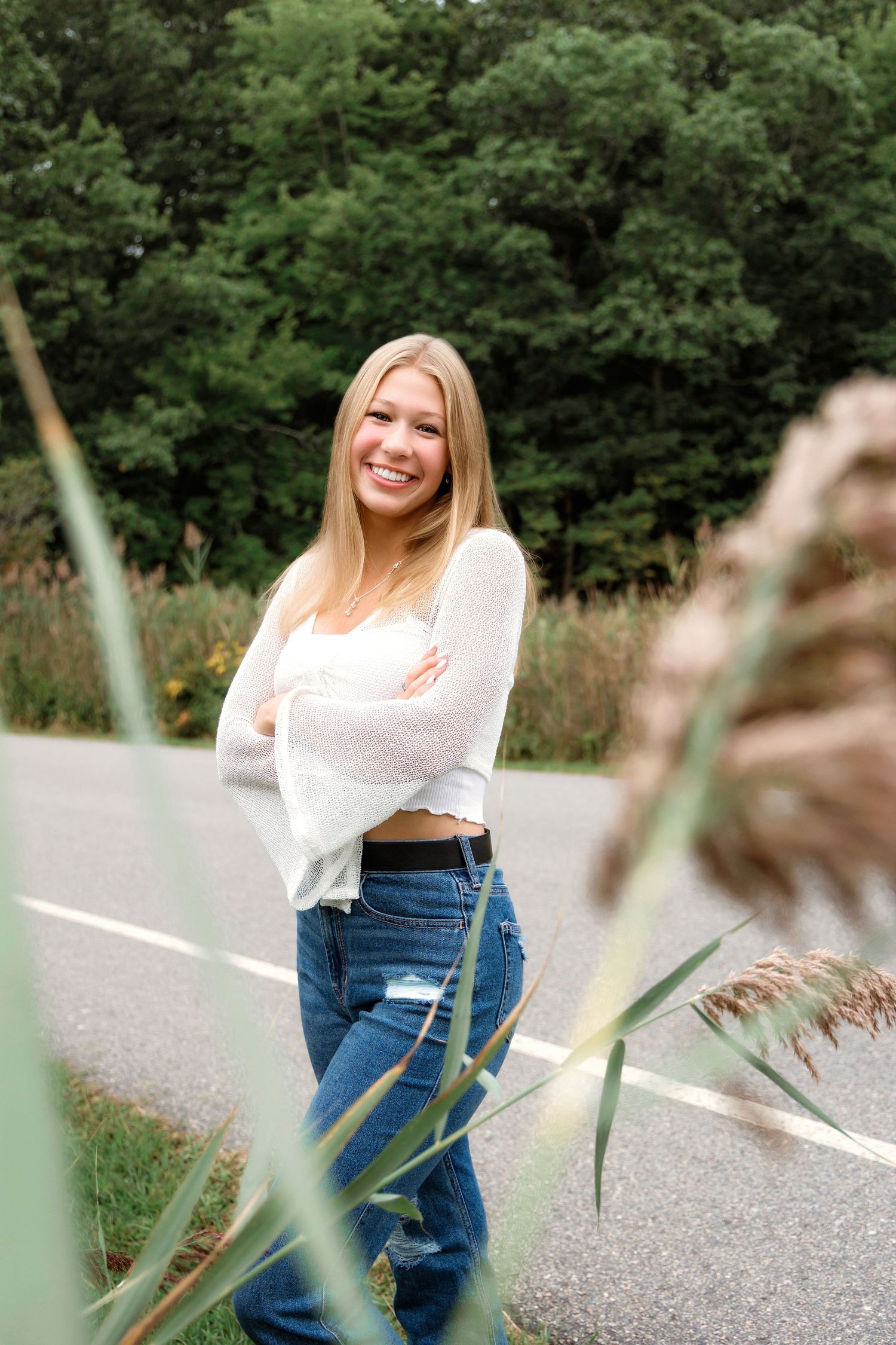 A person in a white top and blue jeans poses on a road with green foliage in the background.
