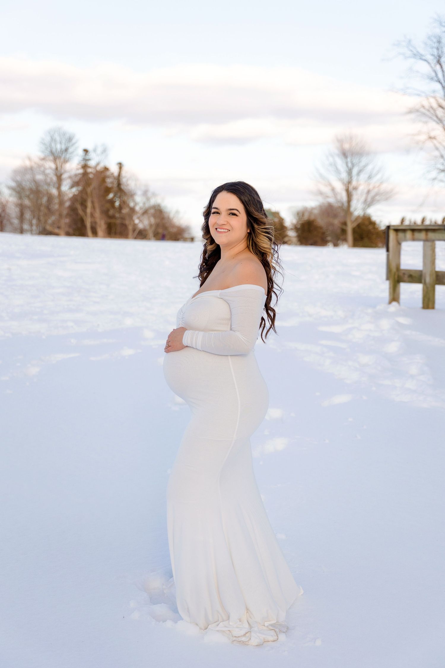 A person in a flowing white dress stands in a snowy winter landscape during a photo session.