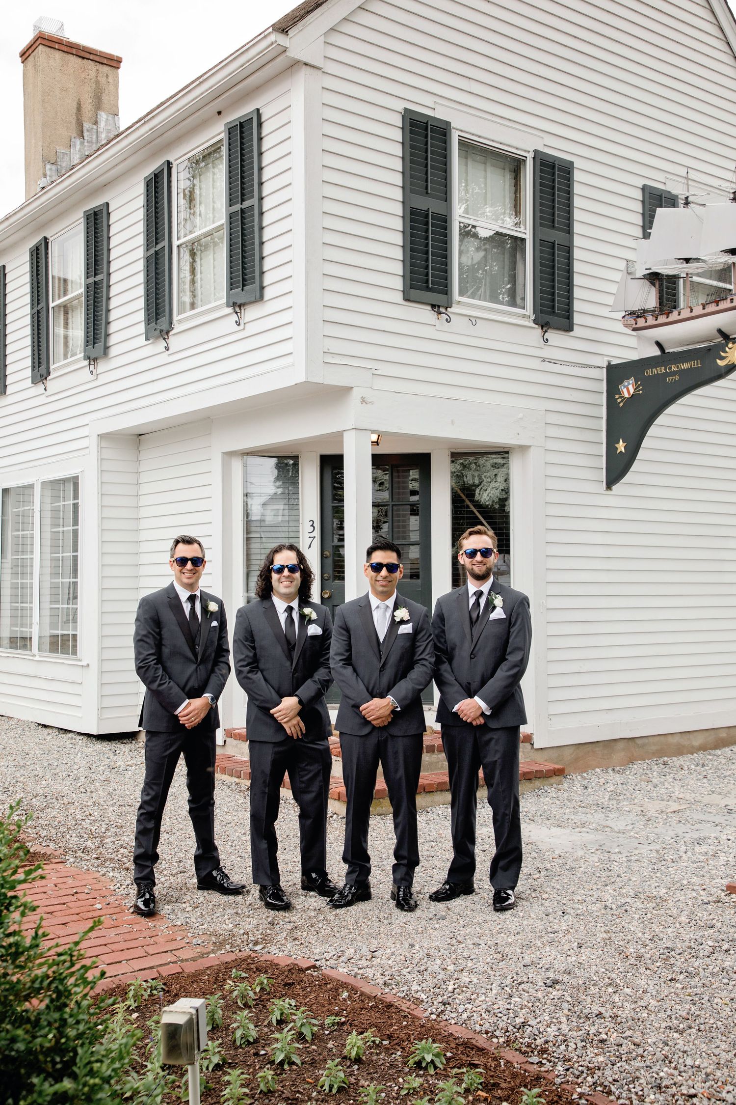 A group of men in black suits and sunglasses pose together on a gravel path next to a white building.