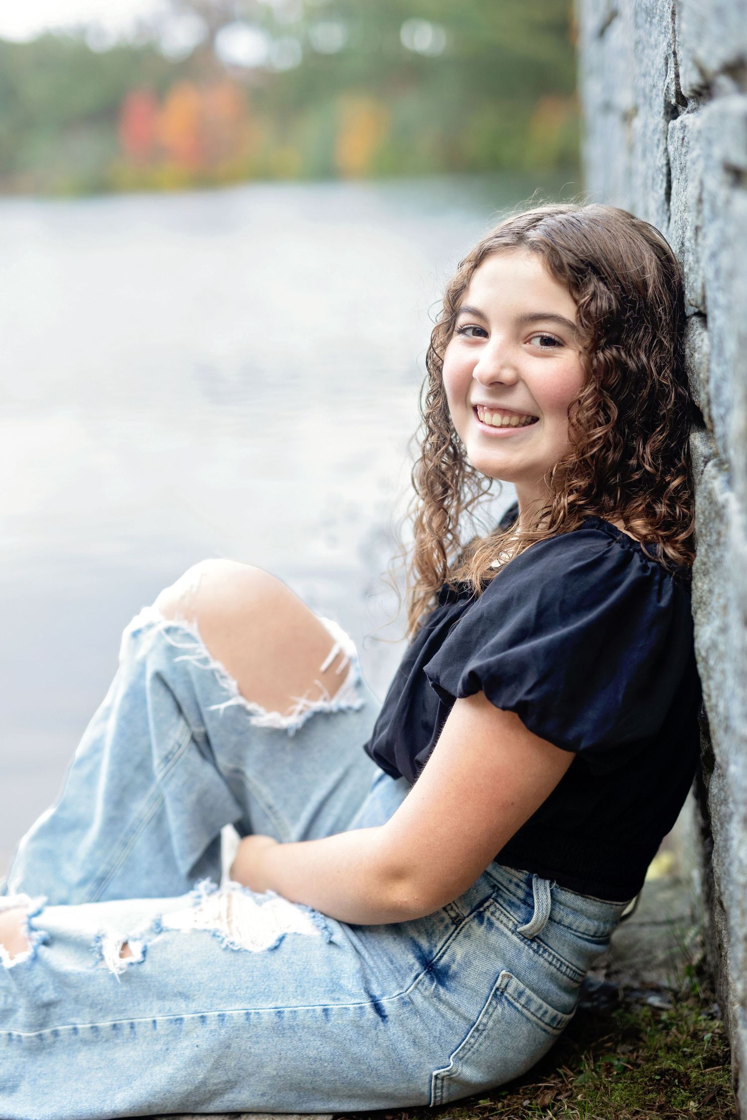 A person in ripped jeans and black top sits against a tree trunk with a natural blurred background.