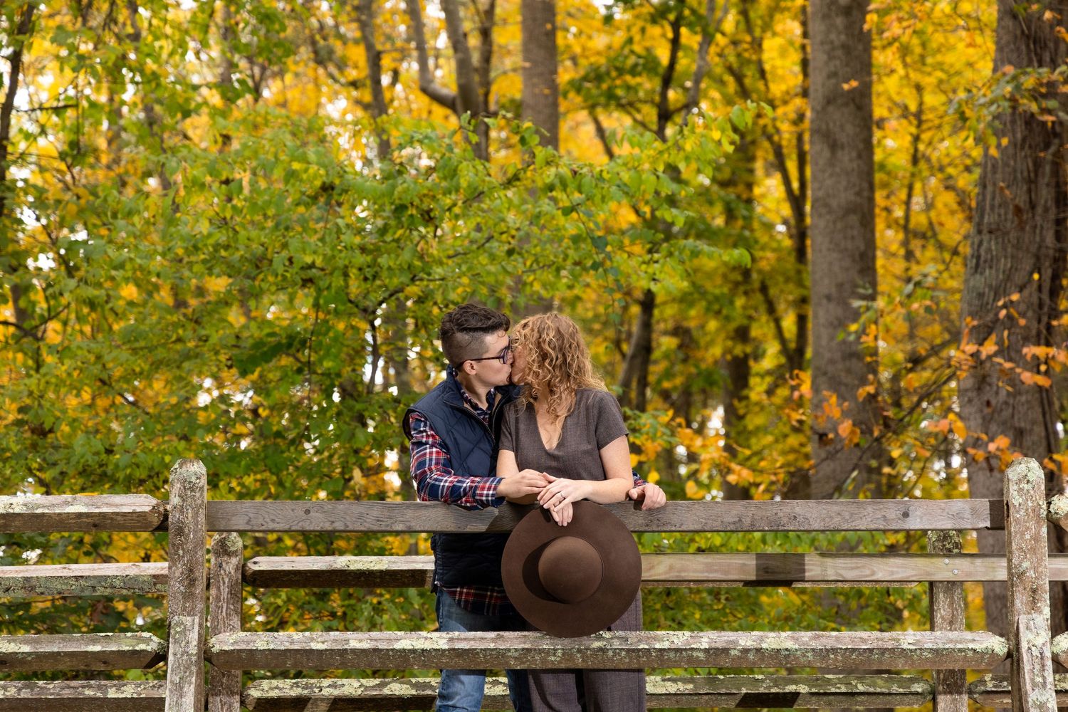 A couple sits close together on a rustic wooden fence surrounded by golden fall foliage.