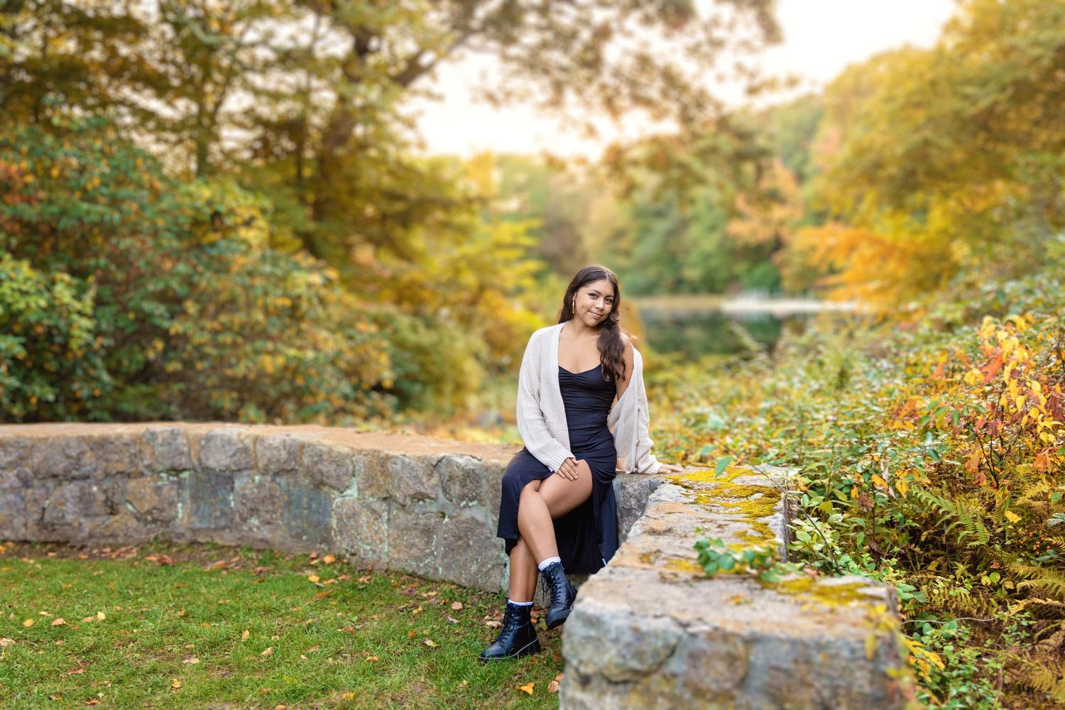 A figure sits on a stone wall surrounded by vibrant autumn foliage during golden hour.