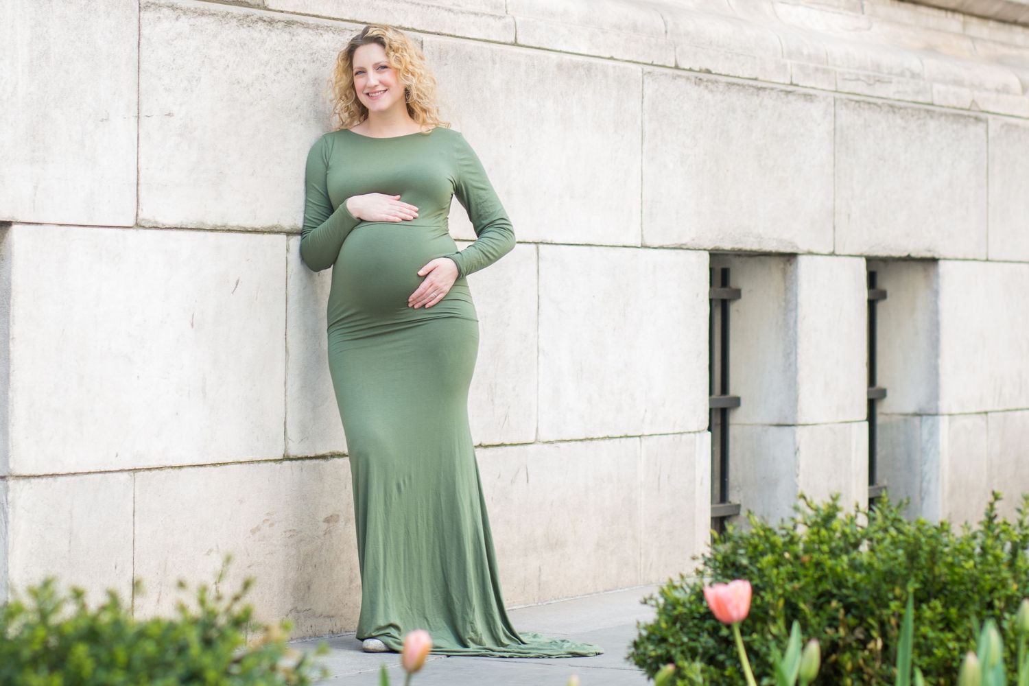 Woman in sage green maternity dress stands against white stone wall with greenery in the background.