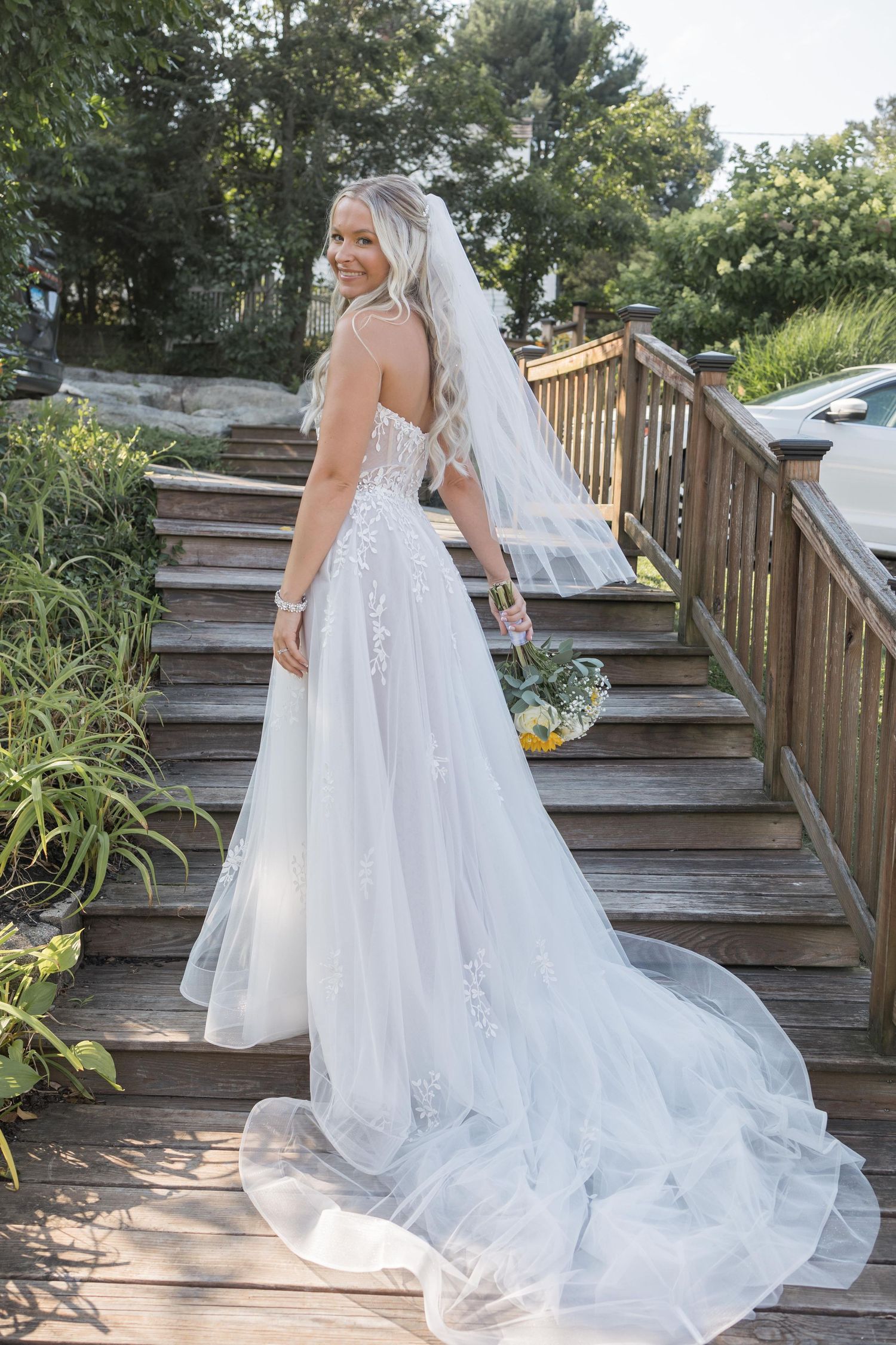 Ethereal wedding dress with flowing tulle train and lace details photographed on rustic wooden deck stairs in natural light.