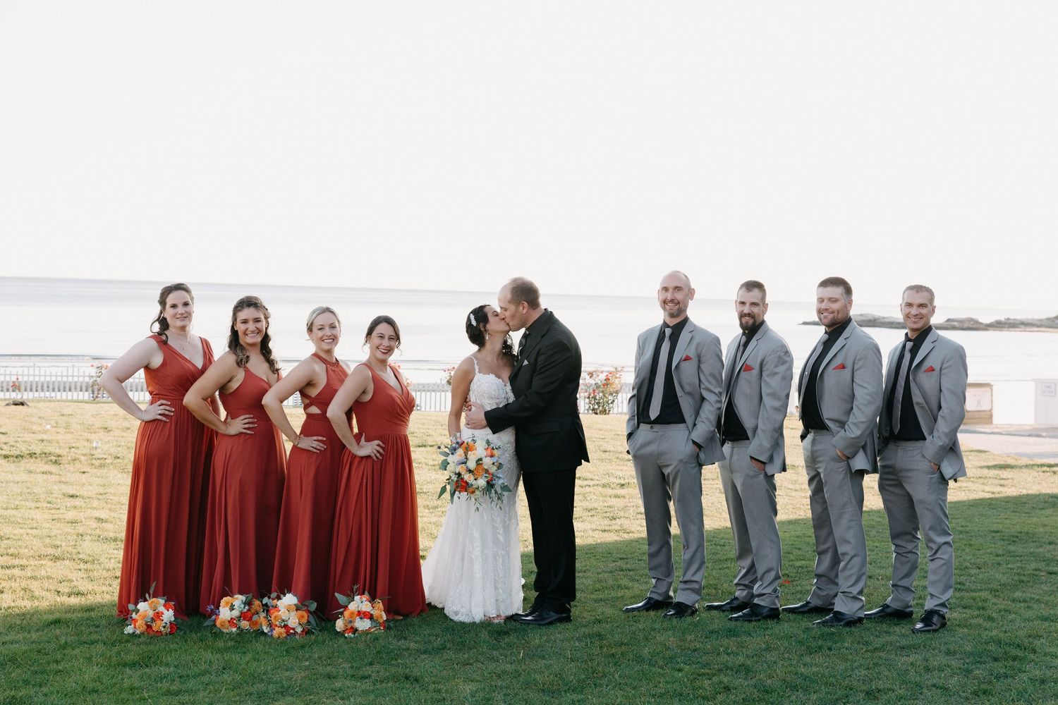 Wedding party lined up on grass with bridesmaids in red and groomsmen in gray.
