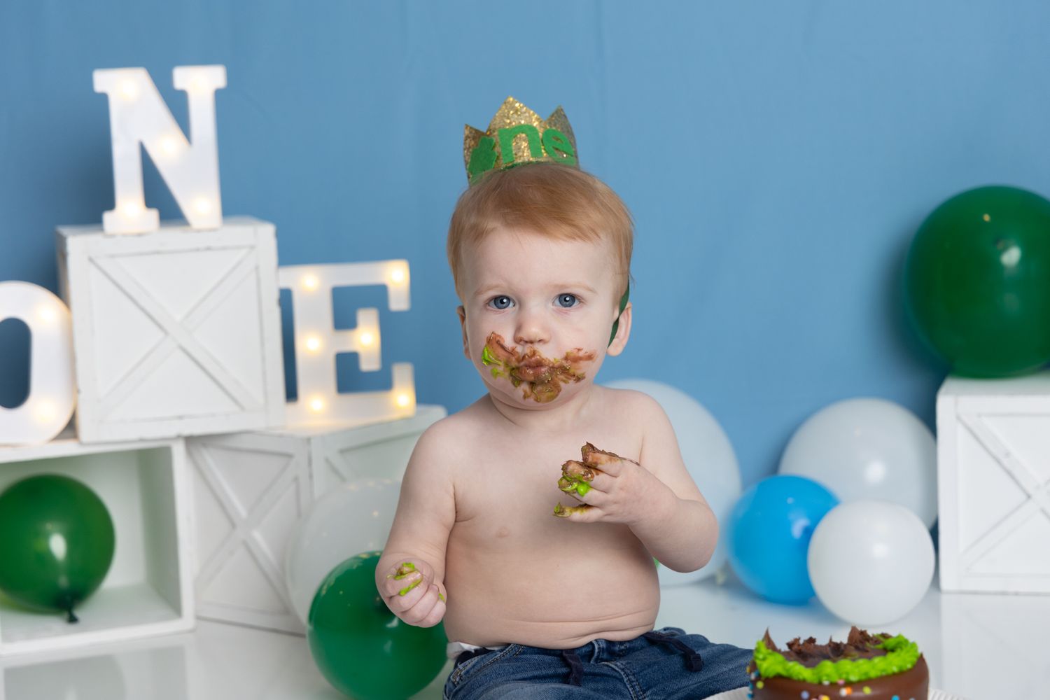 A cake smash photo session with blue and green balloons and white decorative boxes in the background.