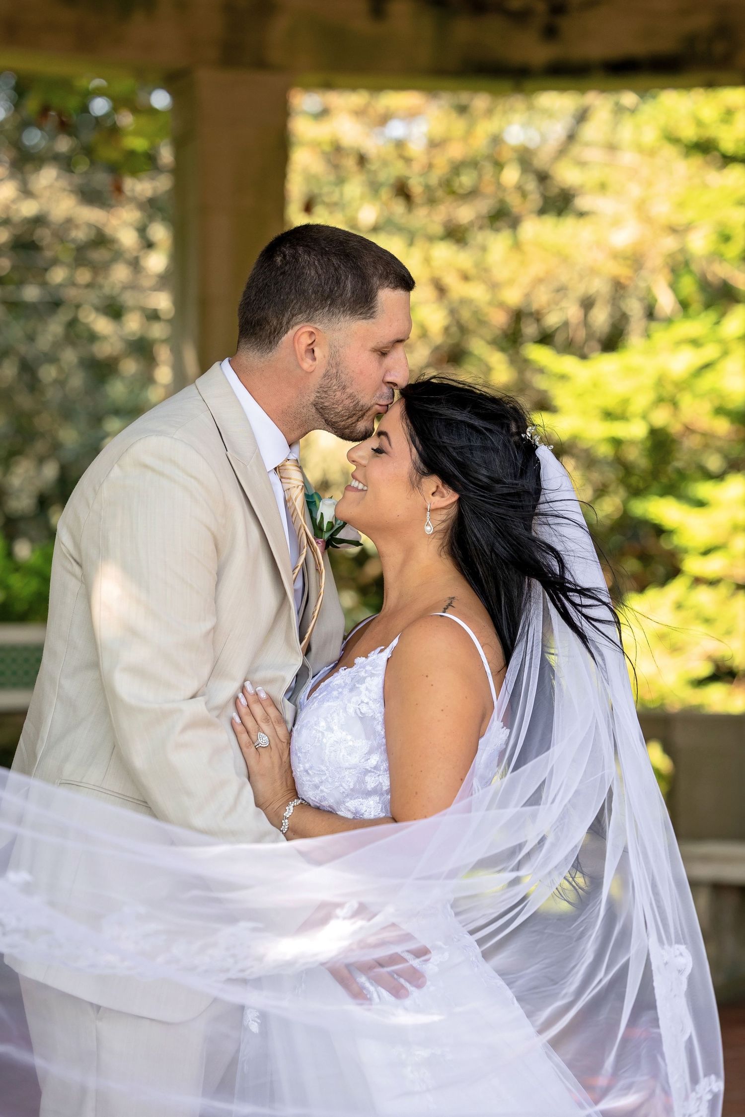 A romantic wedding moment captured with flowing veil and natural sunlit background.