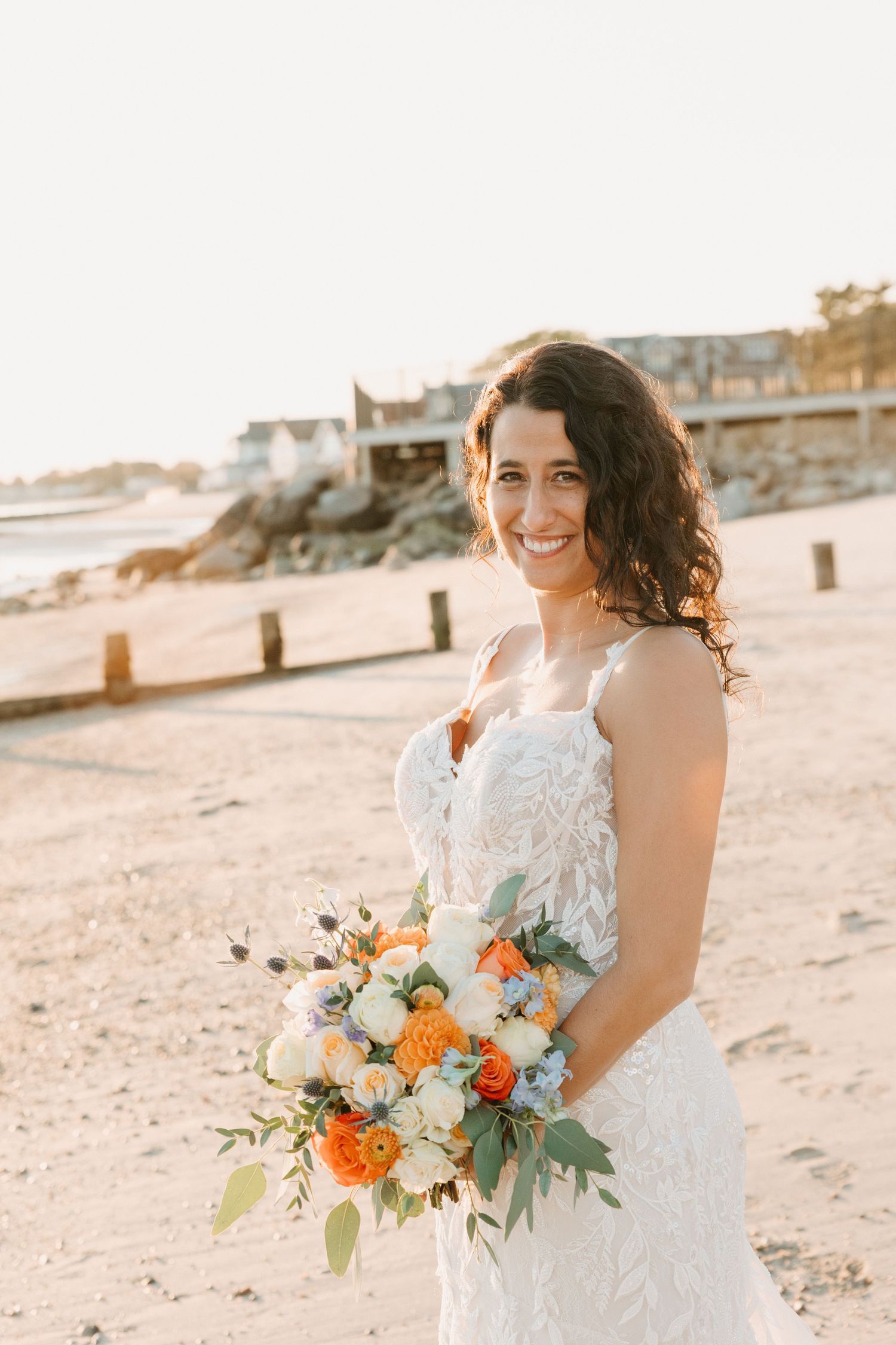 Bride holding orange and white bouquet during sunset beach photoshoot.