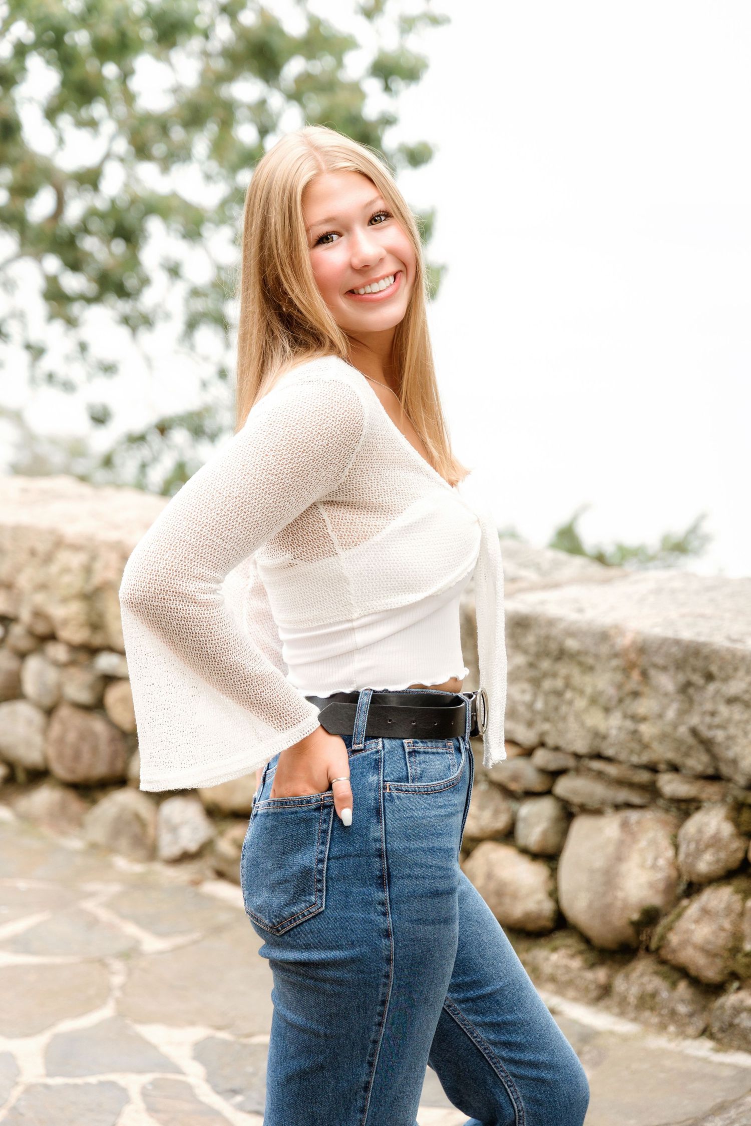 Woman in cream sweater and high-waisted jeans poses casually against stone wall outdoors.