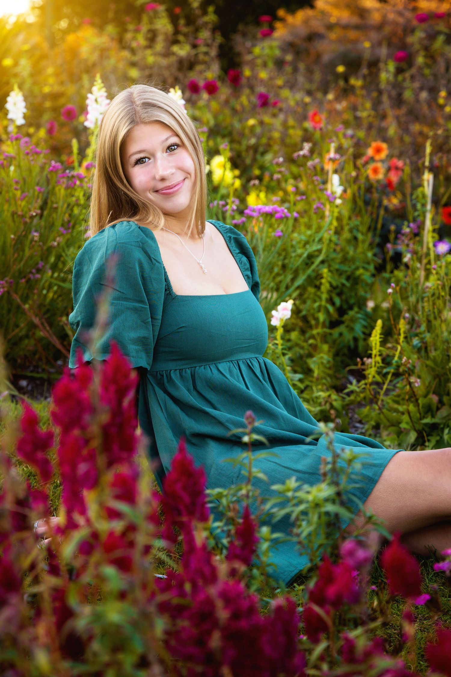 A woman in a teal dress sits among vibrant wildflowers in a garden during golden hour.