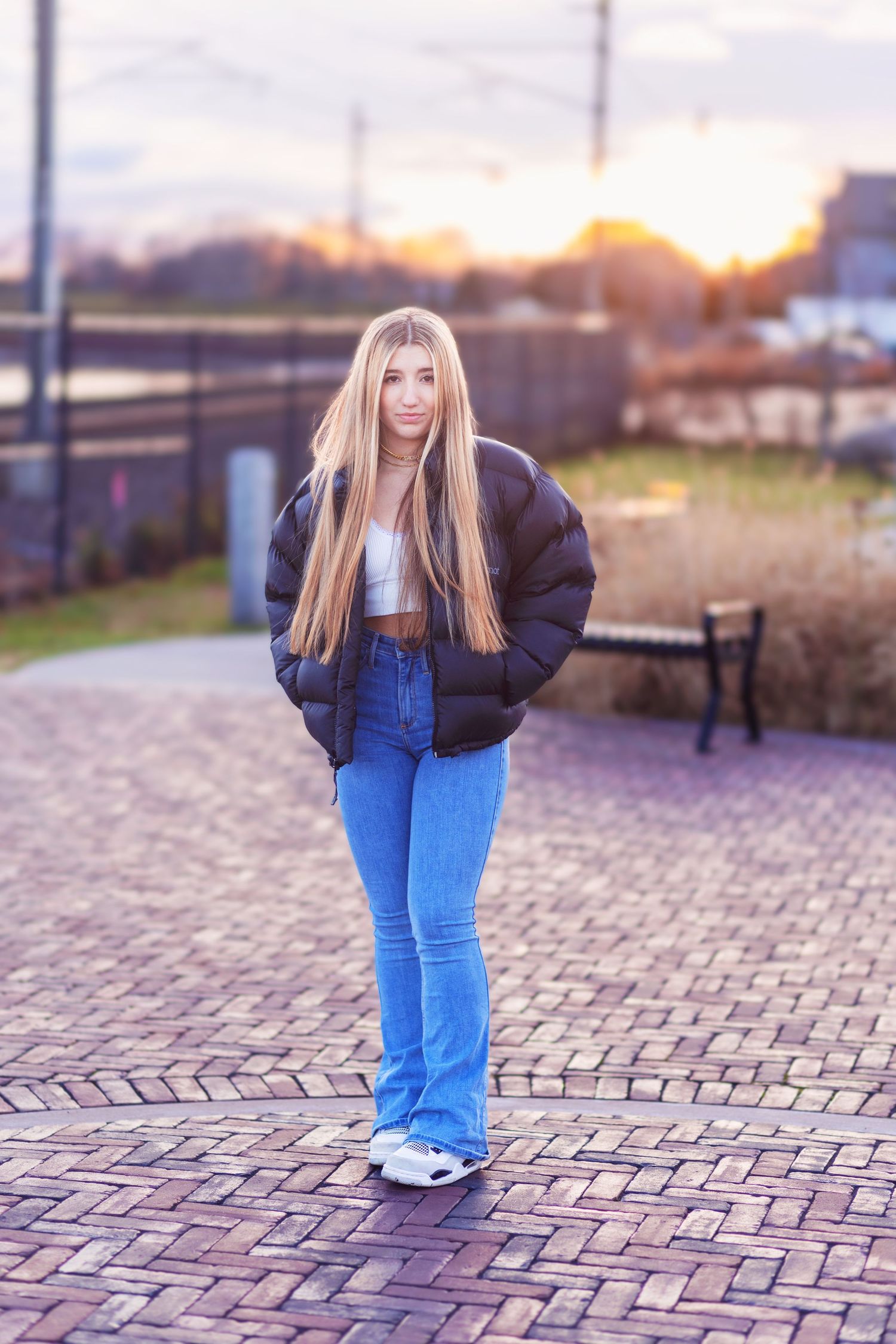 A person in a black puffer jacket and bright blue pants stands on a brick paved walkway at sunset.