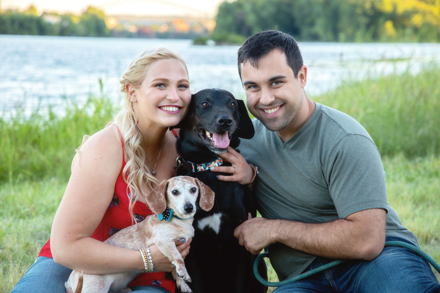 A couple sits outdoors with their black and white dogs during a sunset photo session in a grassy field.