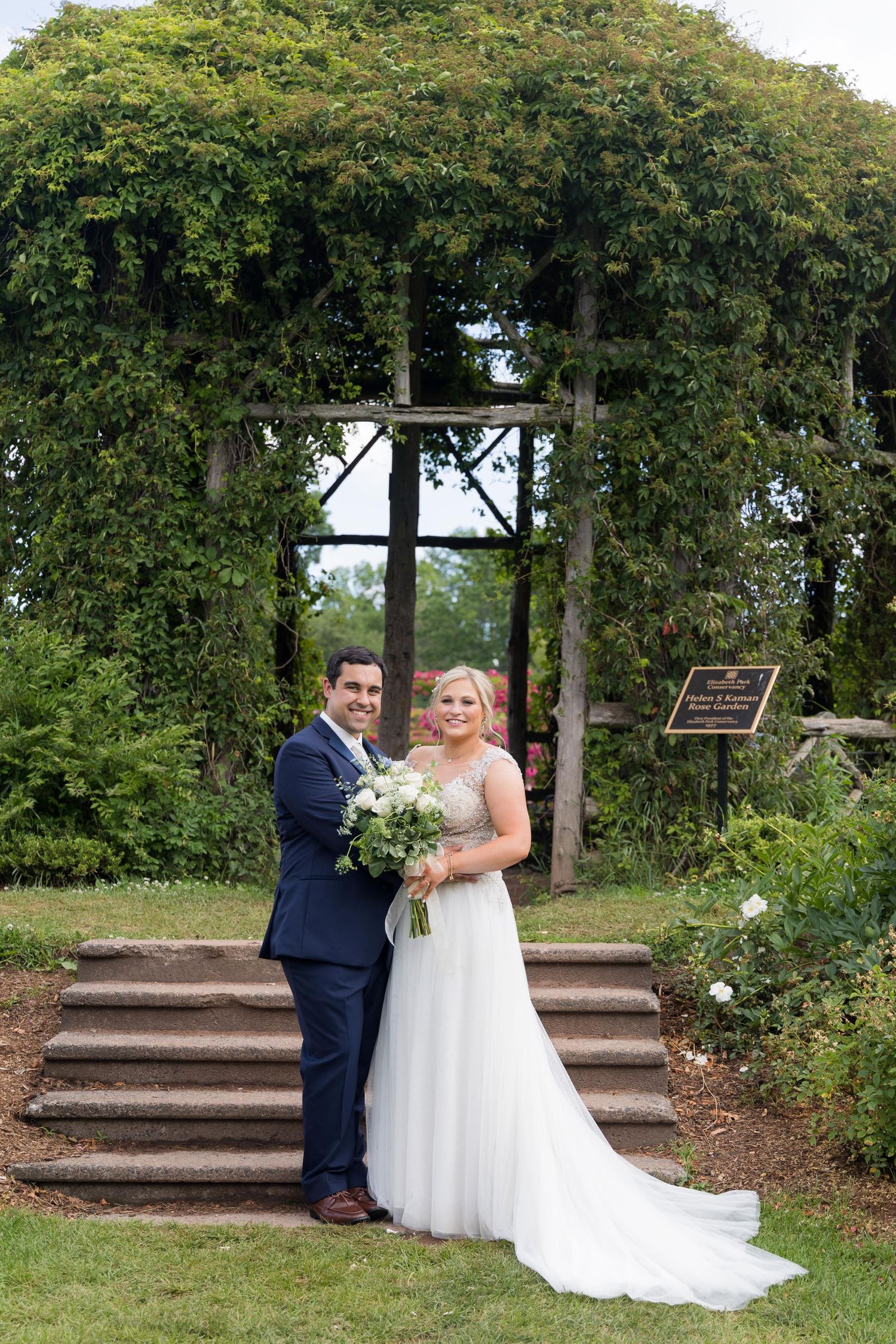 A couple poses together on stone steps surrounded by lush greenery in a romantic garden setting.