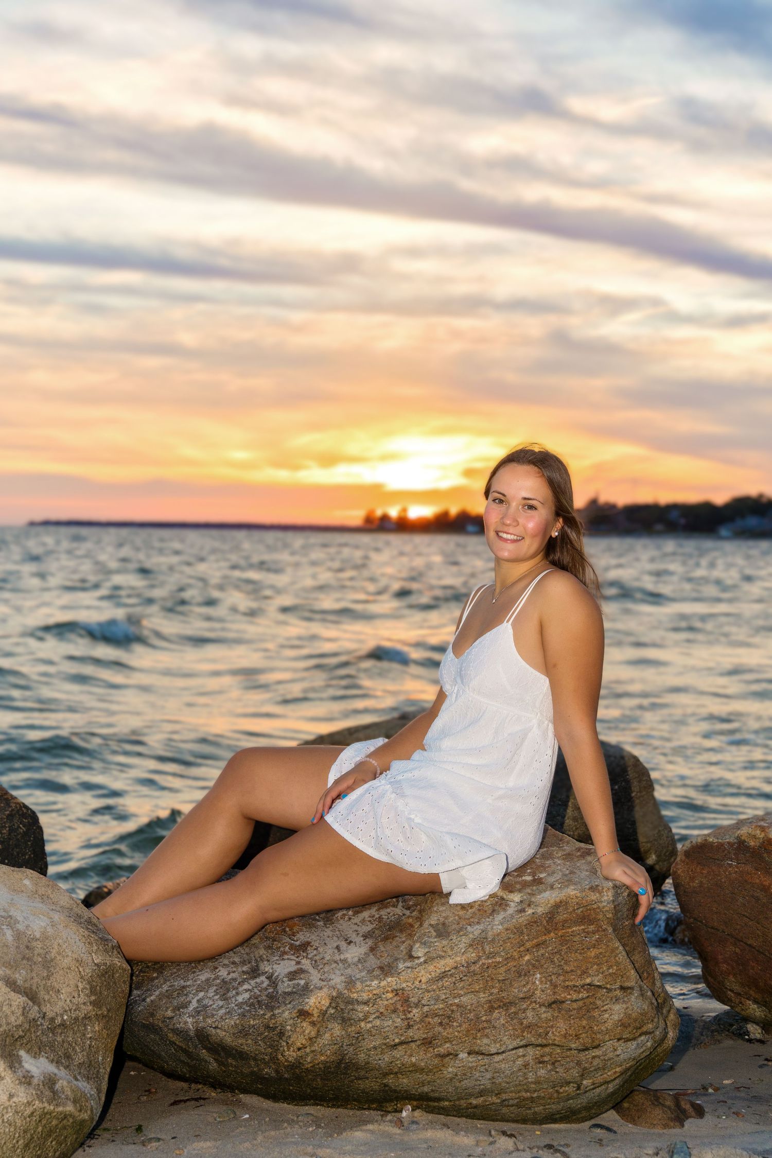 A person in a white dress lounges on coastal rocks at sunset with the ocean and warm sky in the background.