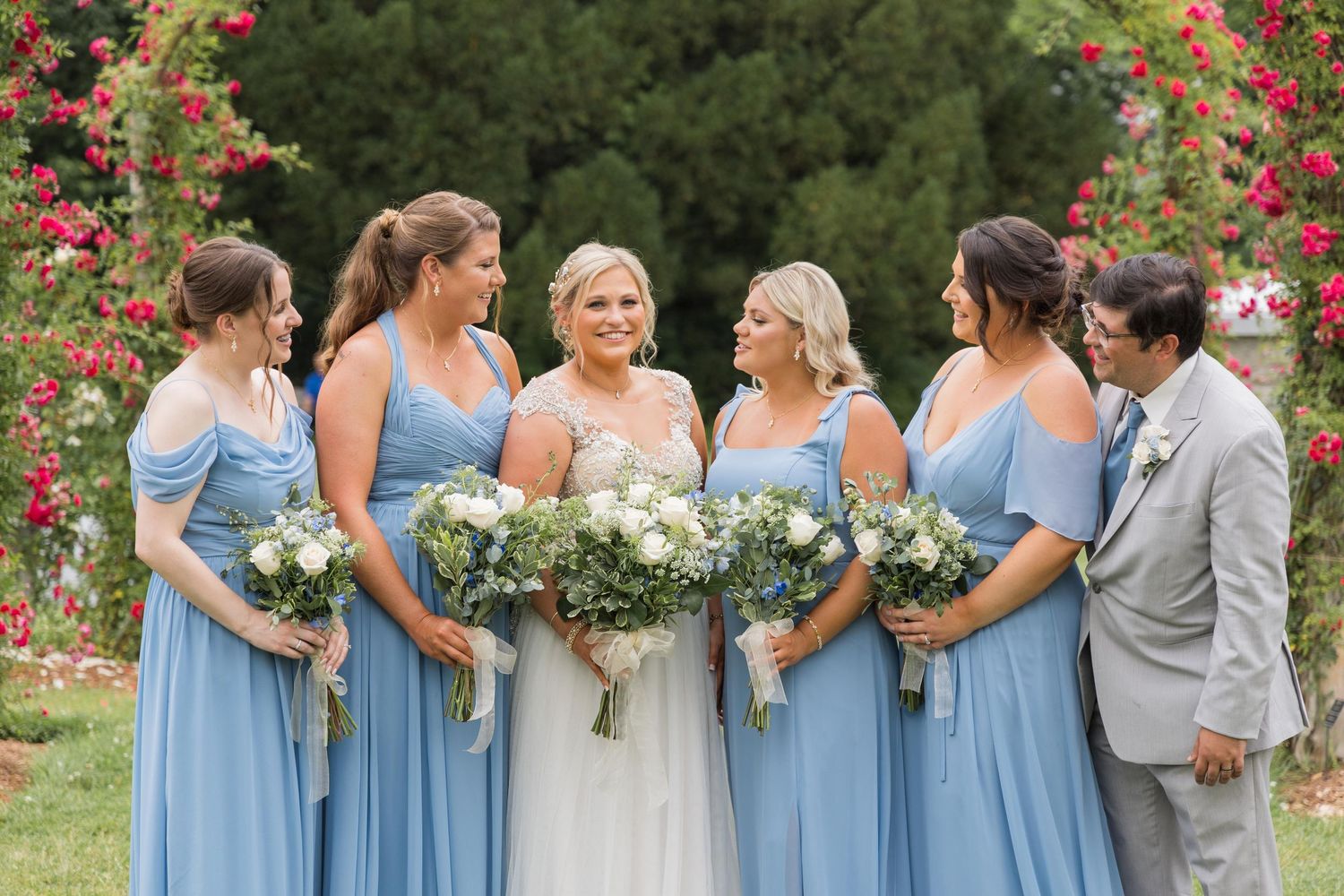Wedding party in light blue bridesmaid dresses and gray suit gather outdoors with white and green bouquets.