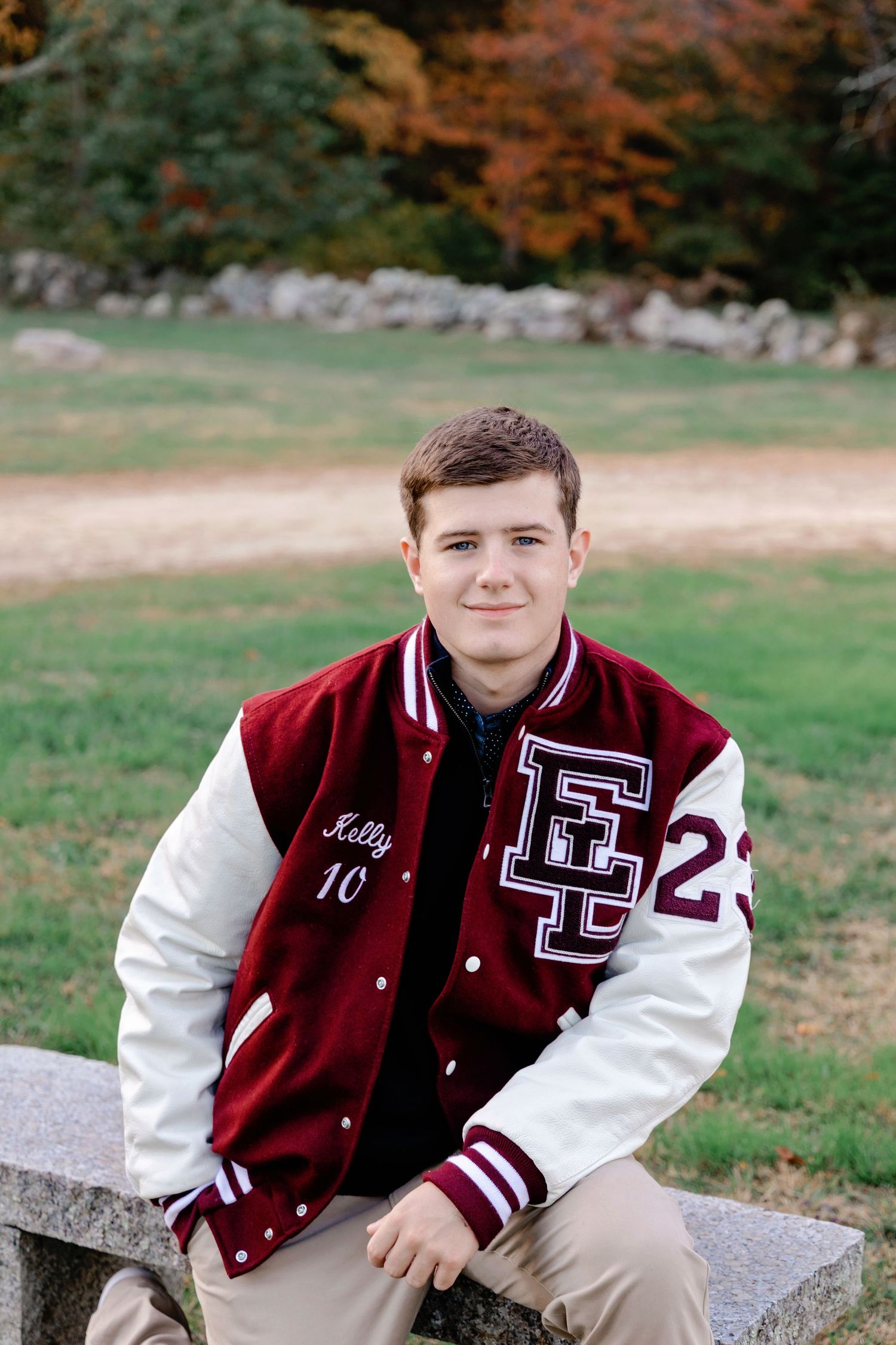 A student in a maroon and white varsity letterman jacket sits outdoors during an autumn photo session.