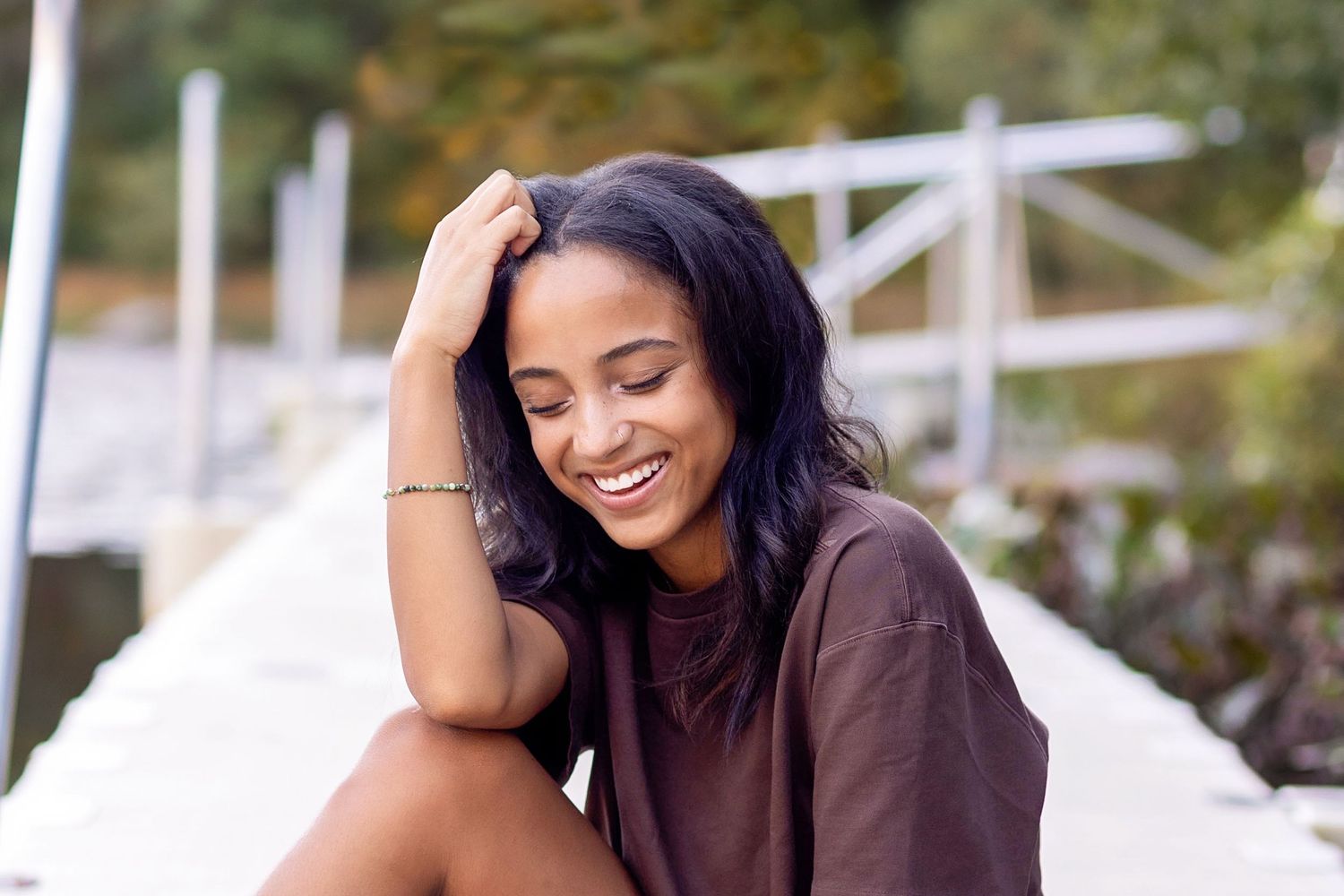 A woman in a brown sweater laughs joyfully while sitting outdoors on a sunny day.