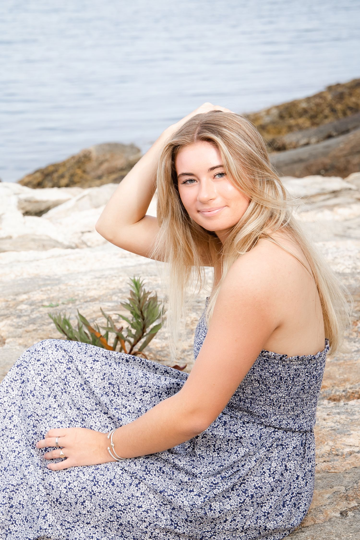 A person in a floral dress poses on rocky coastal terrain at sunset.
