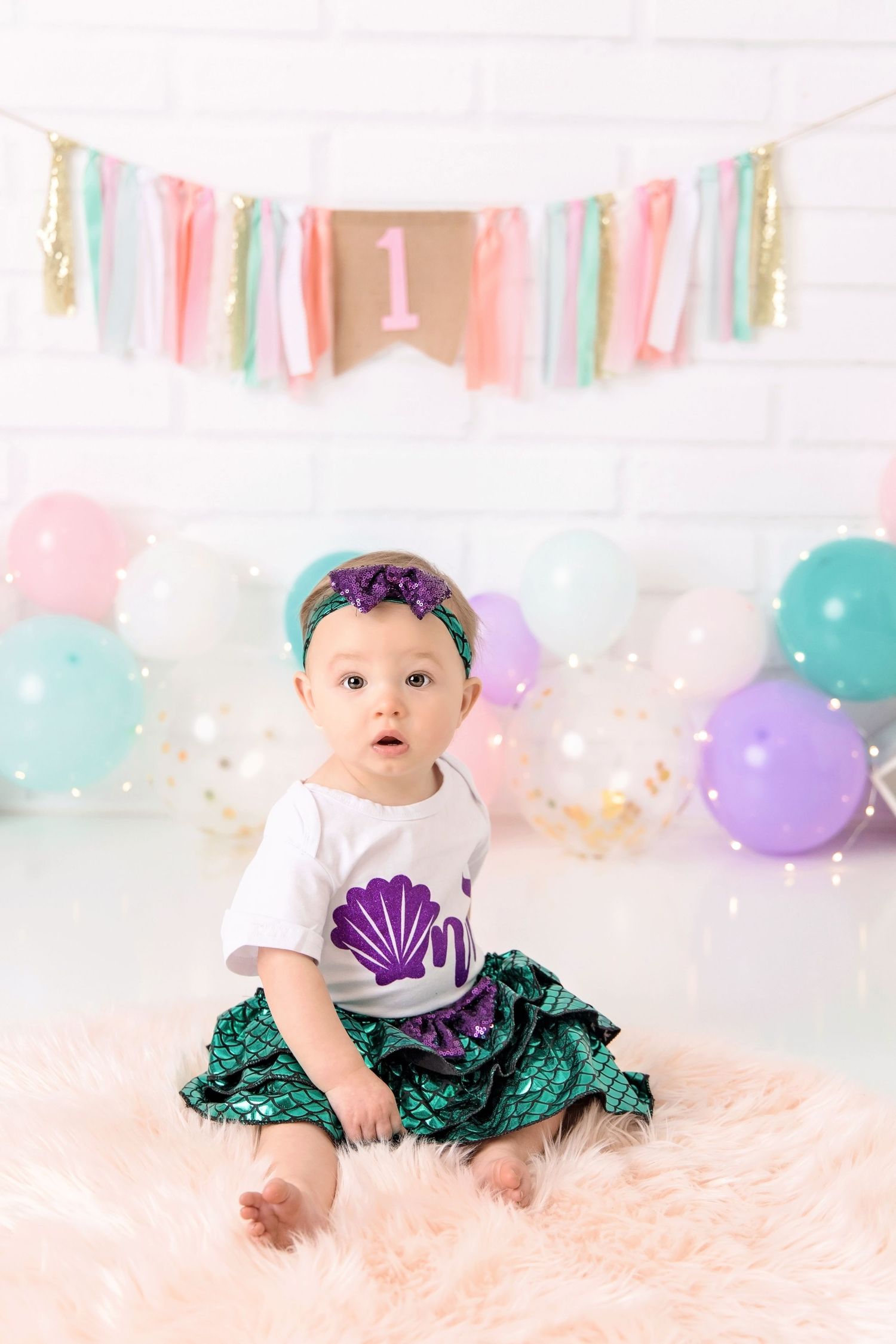 A baby wearing a white shirt and sparkly green tutu sits on a pink rug with pastel balloons and banners in the background.