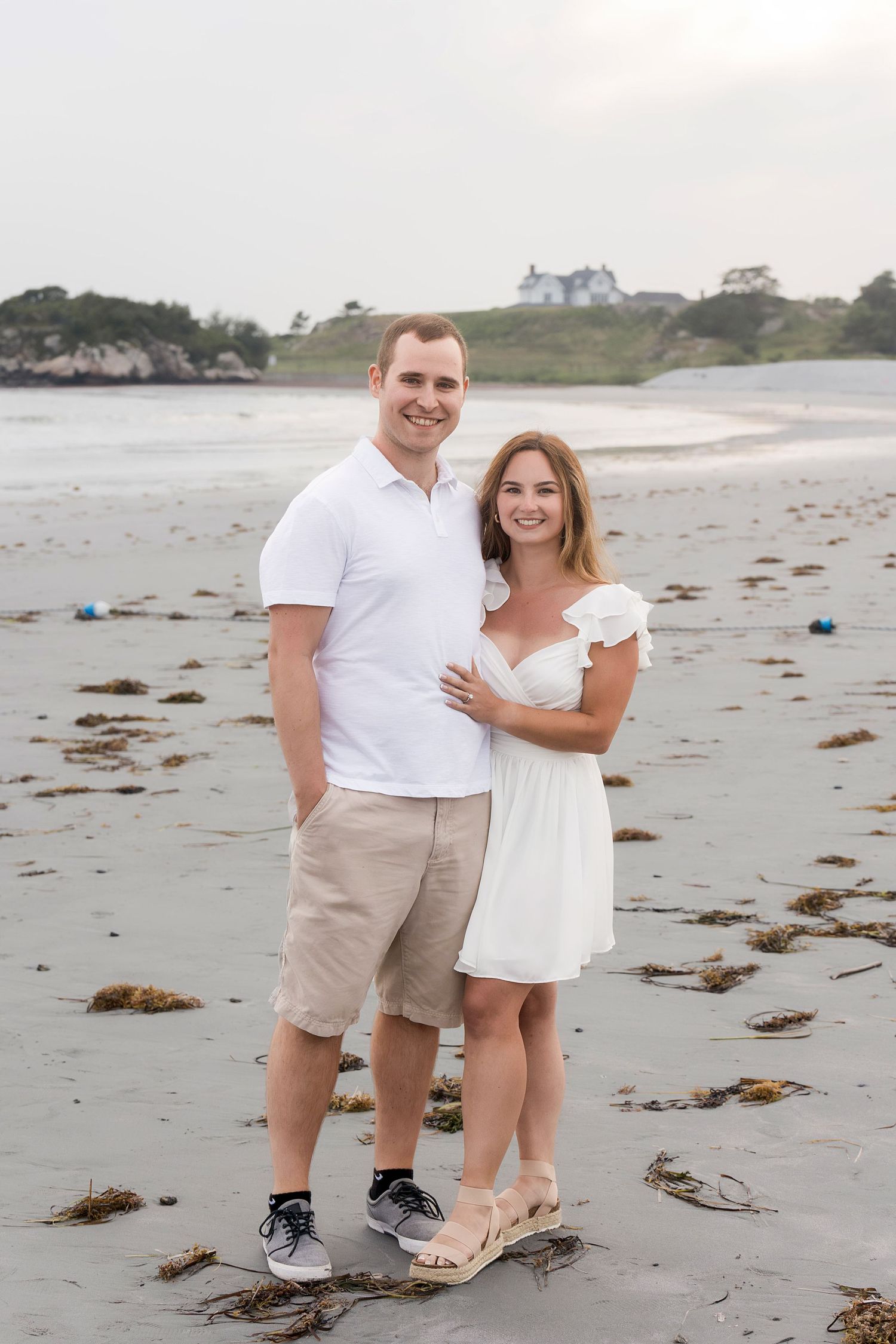 Couple in white and beige outfits share romantic moments during a beach photoshoot at sunset.
